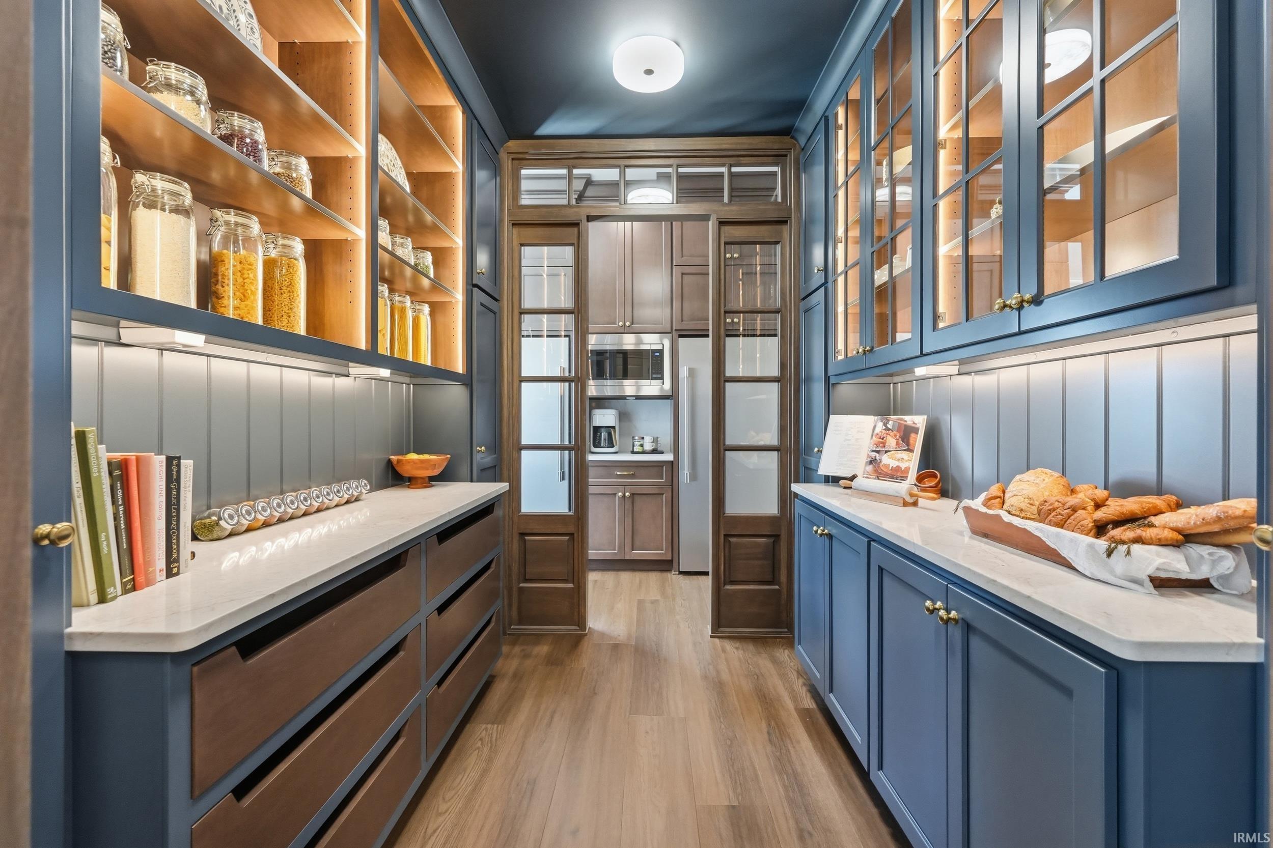 Expansive pantry featuring custom blue cabinetry with brass hardware, illuminated open shelving, wood-finish flooring, and white countertops
