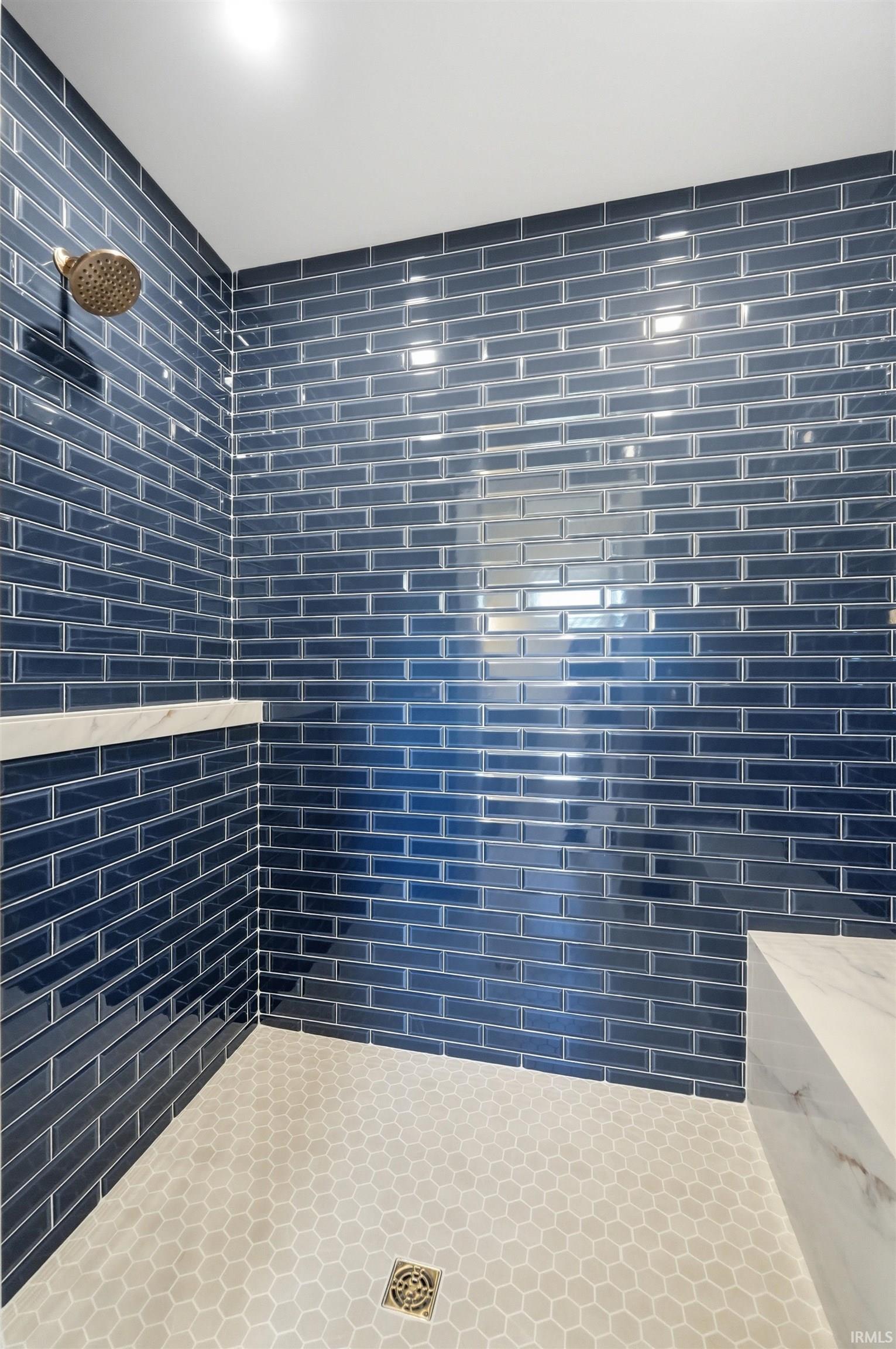 Shower featuring blue subway tile walls, a rain showerhead, a built-in marble-finish bench, a marble-finish accent shelf, and honeycomb floor tiling