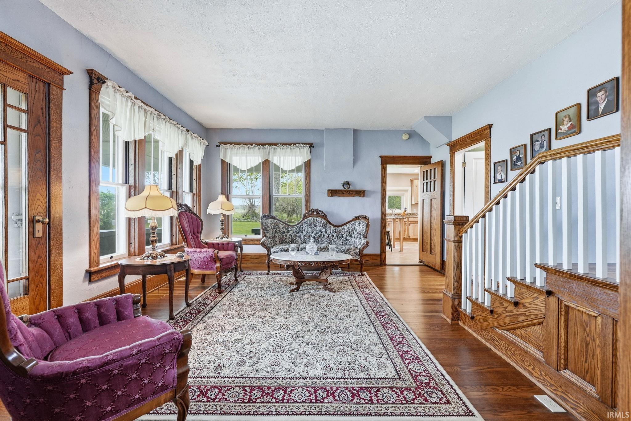 Expansive parlor featuring dark wood-finish flooring, numerous windows with white trim, a detailed staircase with wood treads and white balusters, and substantial wood-frame doorways