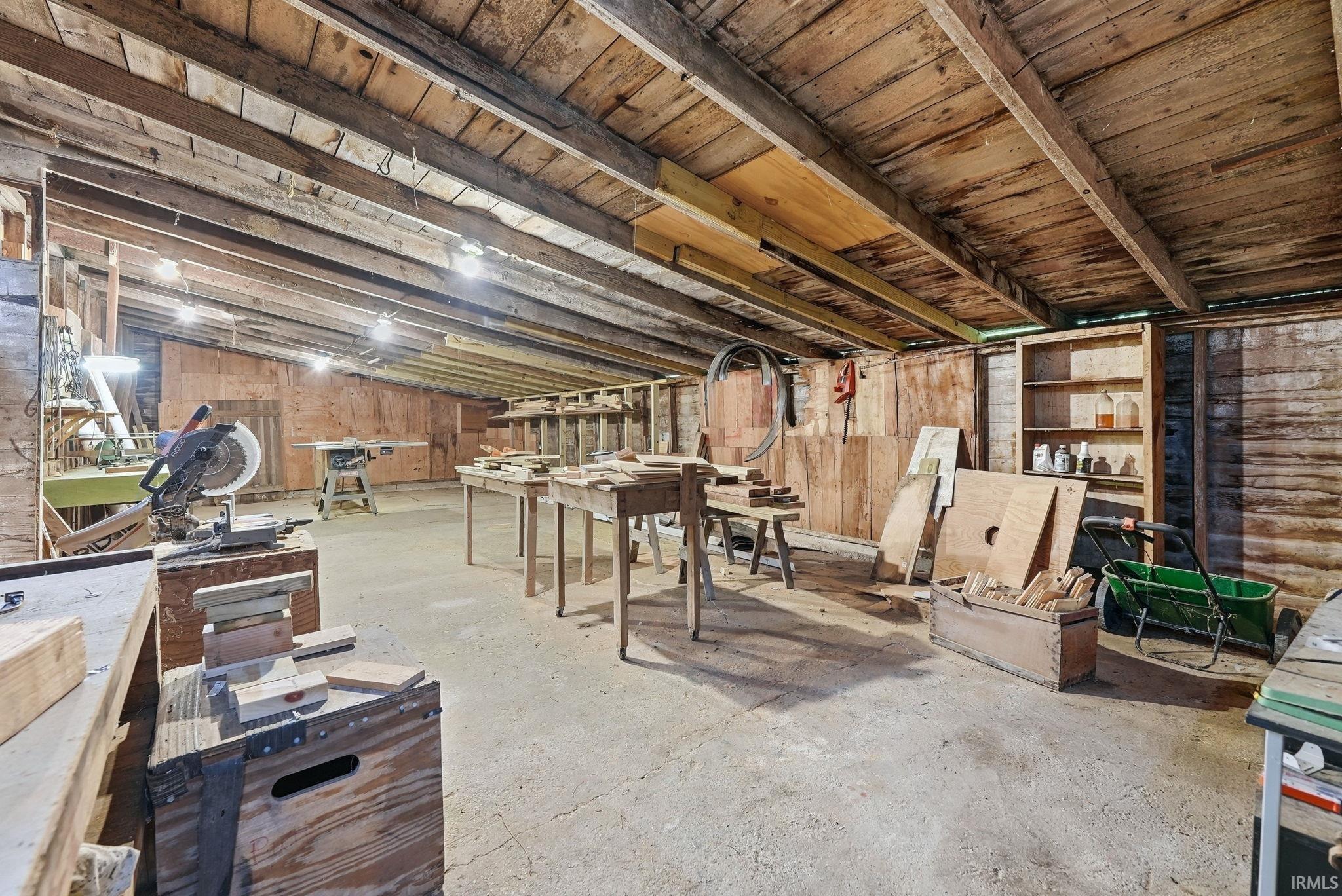 Open workshop space featuring exposed wood beam ceiling, concrete flooring, built-in shelving, and several workbenches