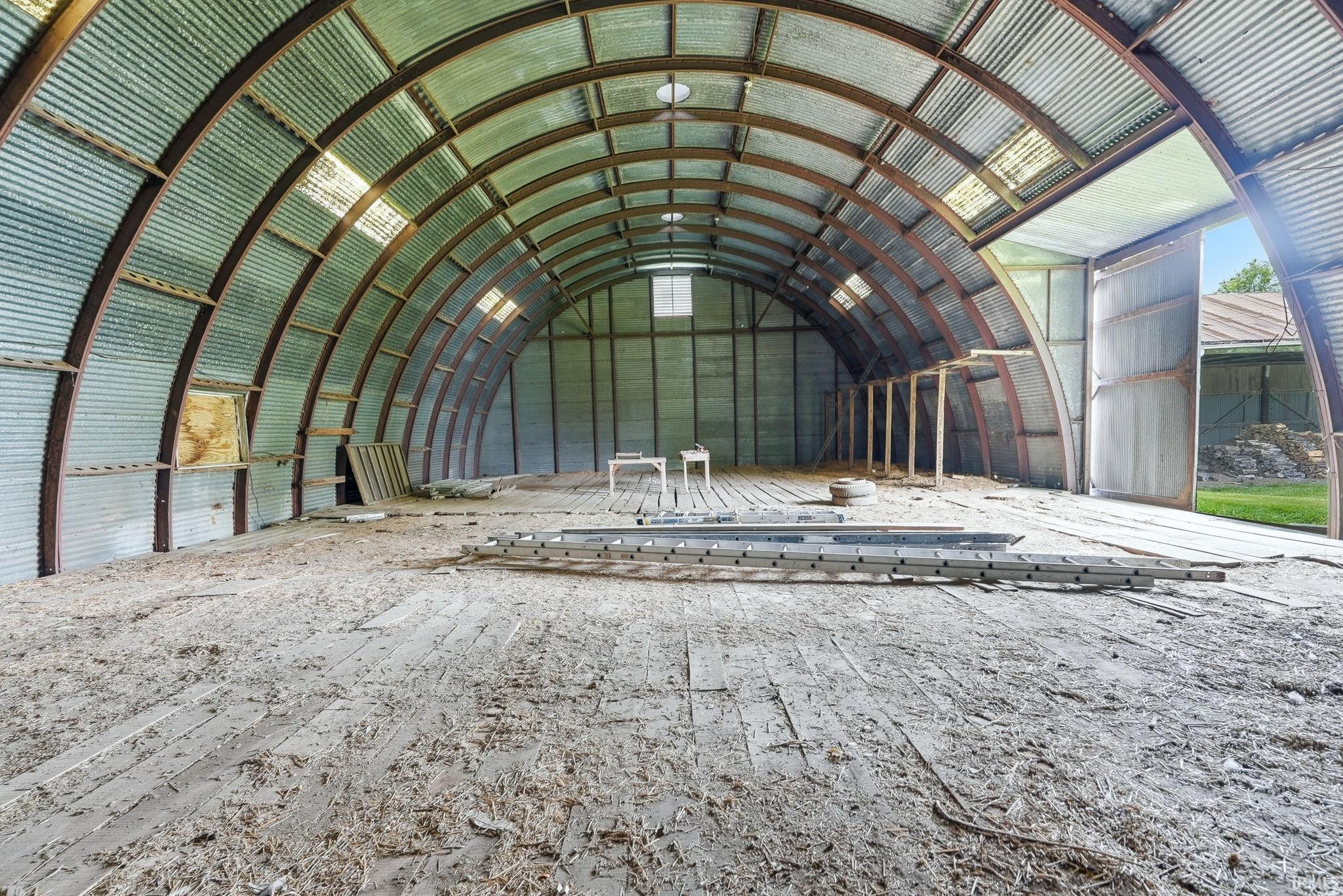 Expansive clear-span structure featuring a corrugated metal roof, exposed arching metal trusses, wood plank flooring, and multiple translucent roof panels providing natural light