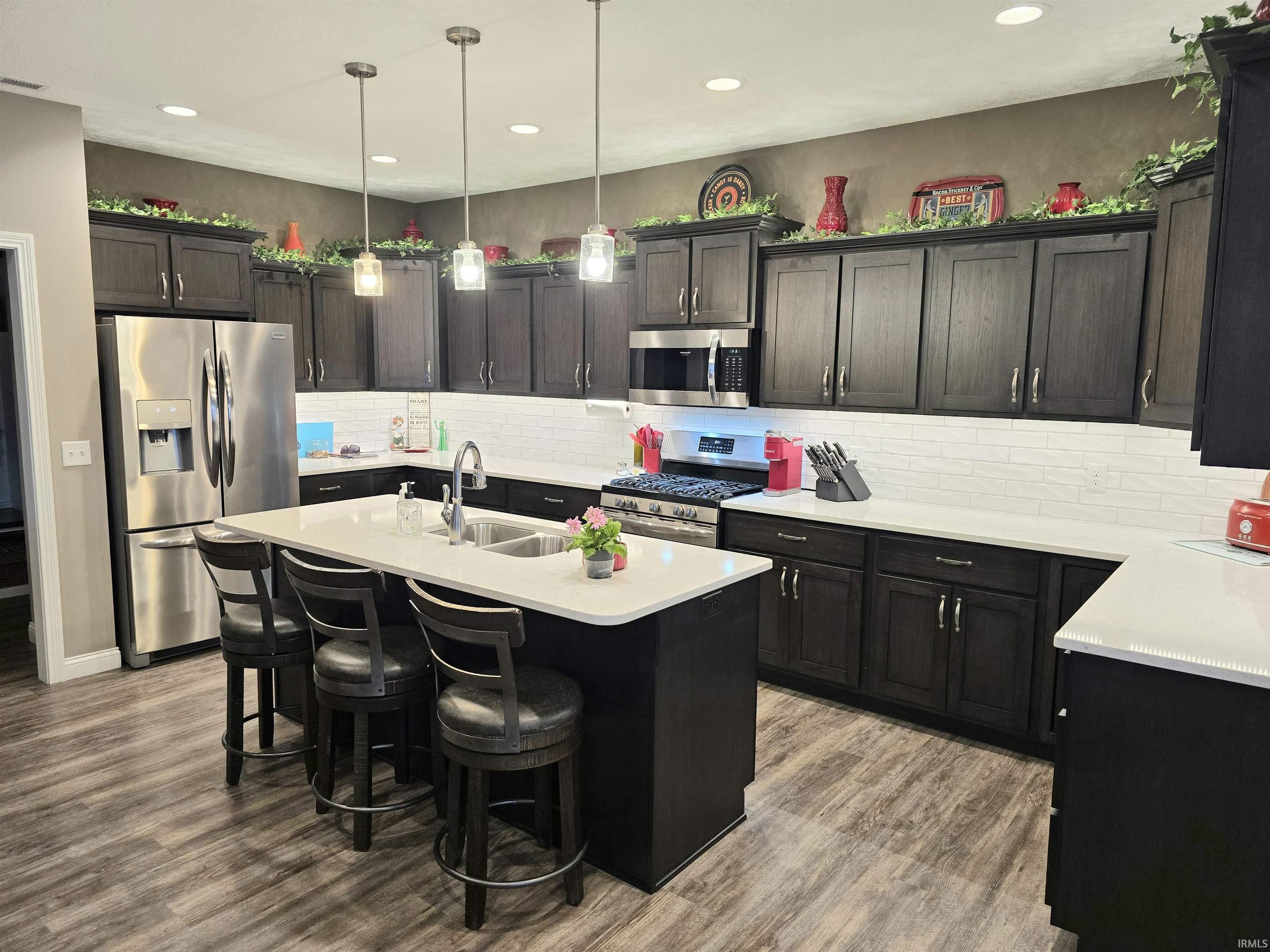 Kitchen featuring dark wood-finish cabinetry, stainless steel appliances, a white subway tile backsplash, light-toned countertops, and LVP flooring.