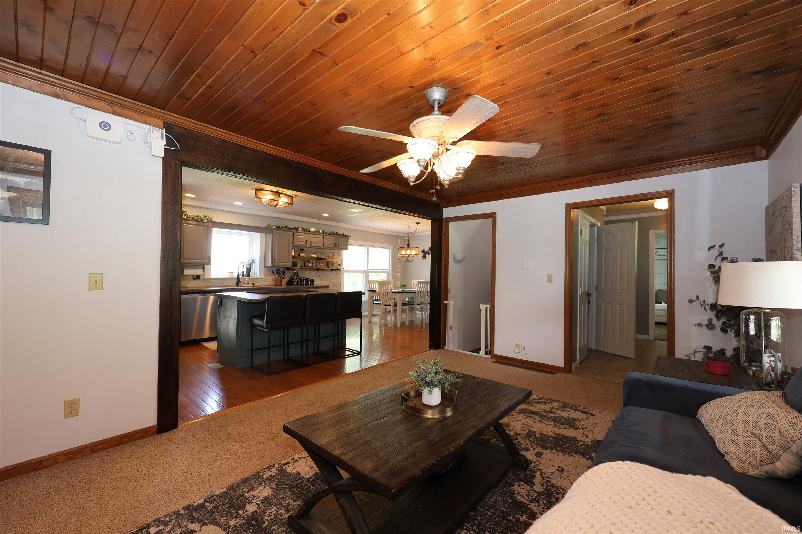 Living area featuring wood-paneled ceiling with a ceiling fan, carpeted flooring, and a large archway opening into the kitchen