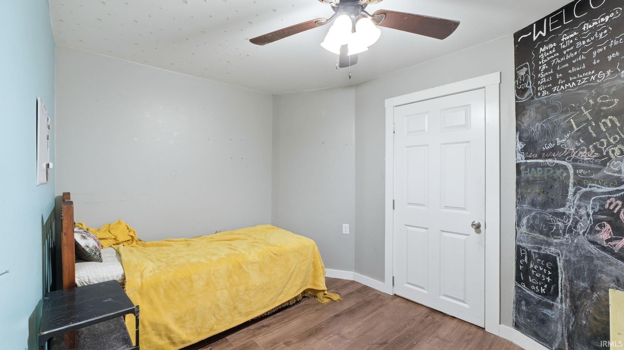Interior room featuring wood-finish flooring, a white six-panel door, a ceiling fan with integrated lighting, and a prominent chalkboard accent wall