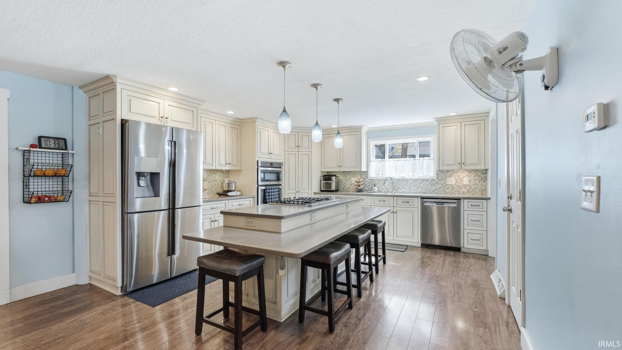 Kitchen featuring an expansive center island with seating, pendant lighting, stainless steel appliances, wood-finish flooring, and light-toned cabinetry