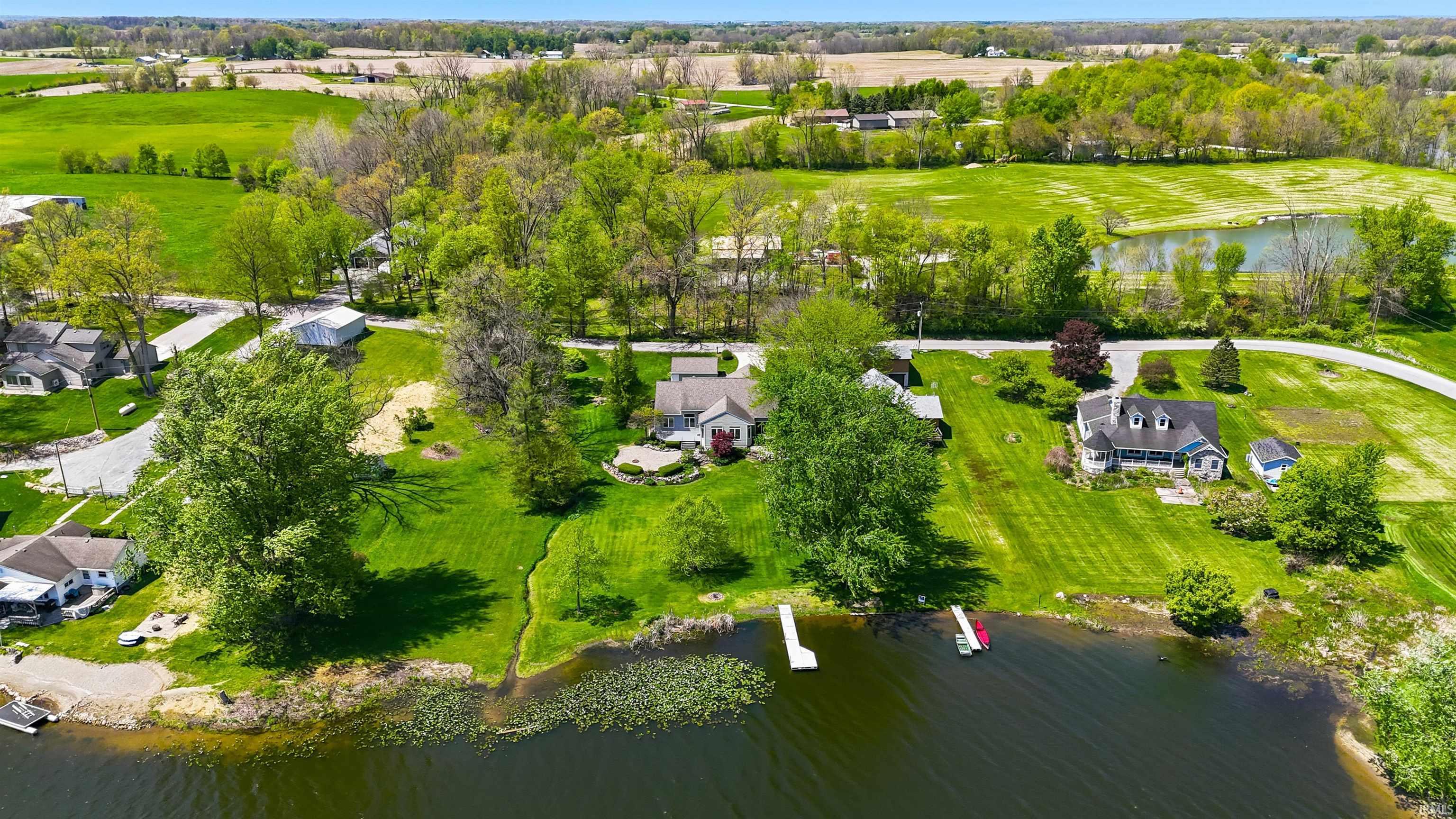 Waterfront property featuring a private dock, expansive green lawn, mature trees, and a gravel shoreline