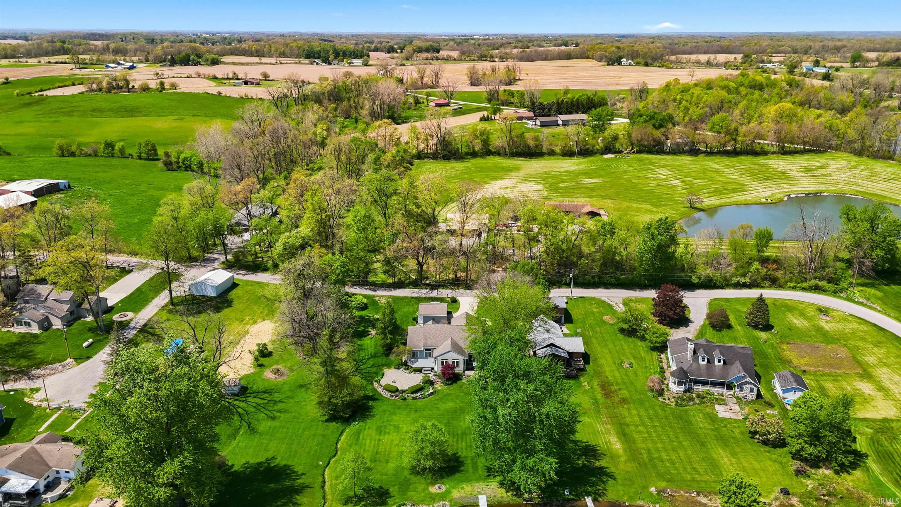 Expansive green spaces and mature trees surround residential properties, featuring a visible pond and distant agricultural fields