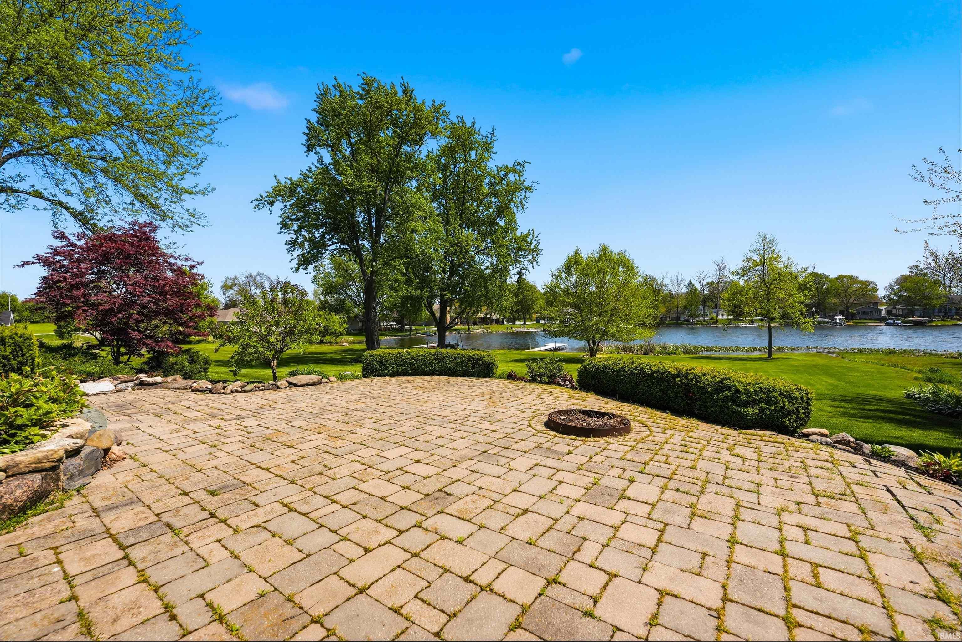Paver patio featuring a built-in fire pit, surrounded by lush green lawn and mature trees, with a body of water visible in the background
