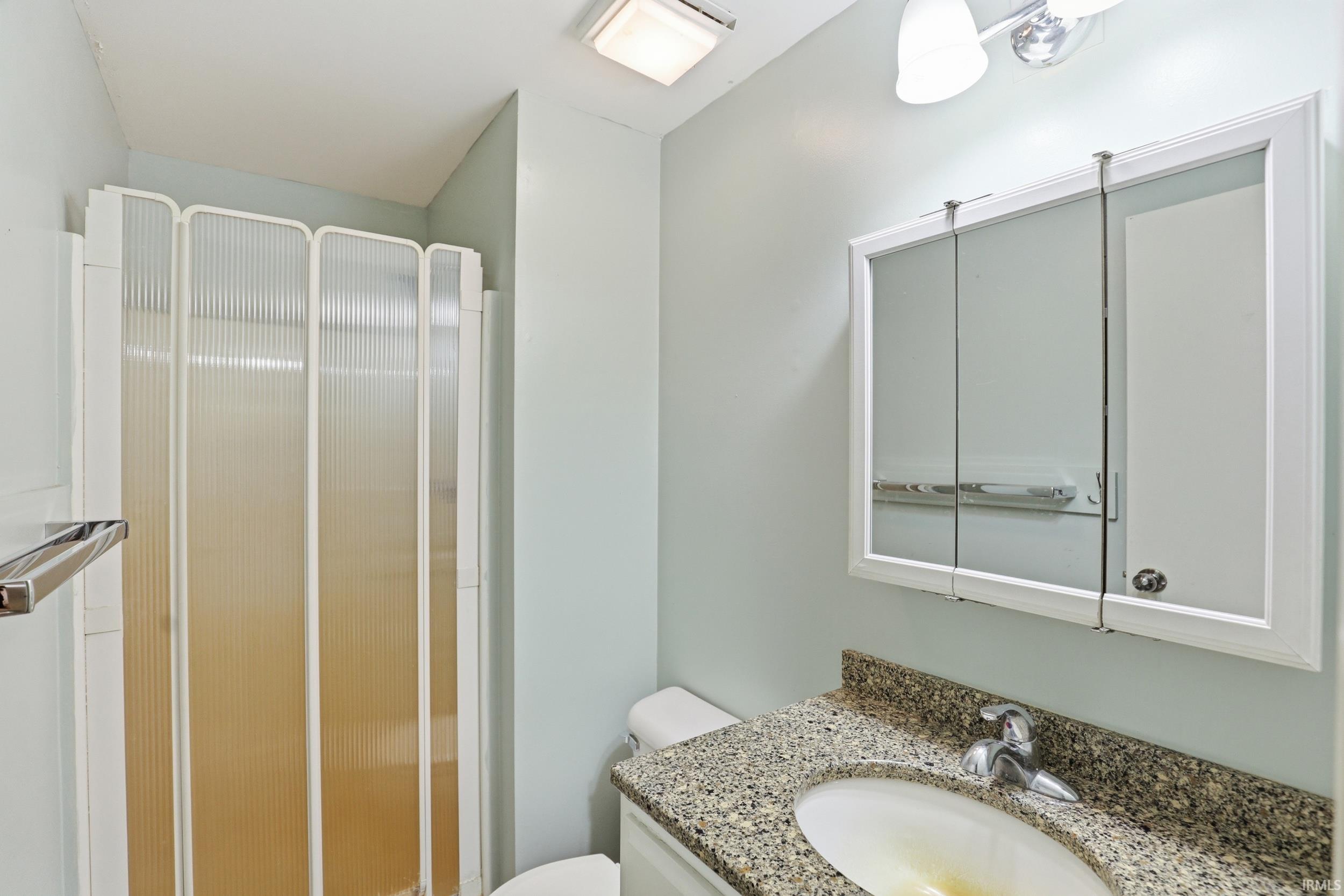 Bathroom featuring a shower enclosure with translucent accordion doors, a vanity with a speckled countertop and integrated sink, a mirrored medicine cabinet, and a two-bulb wall sconce