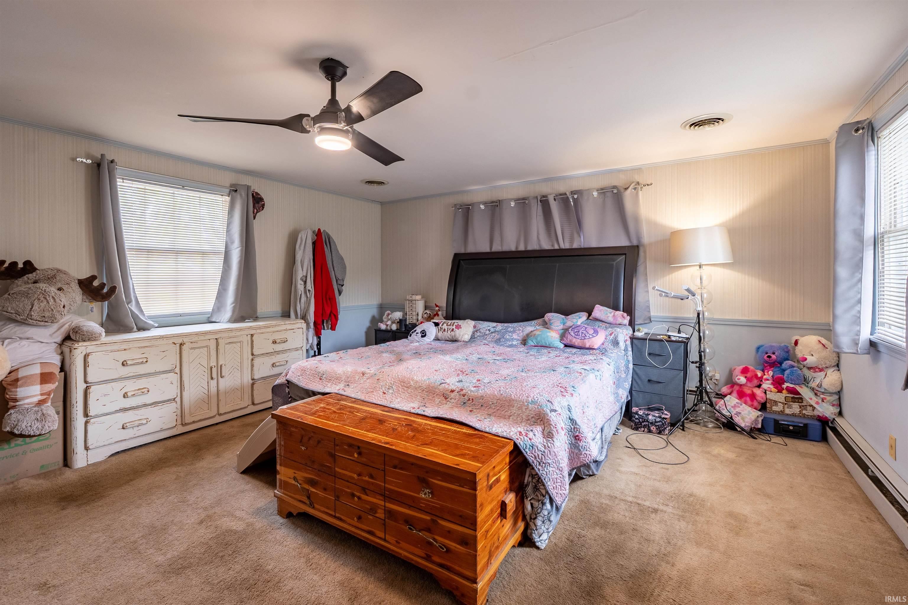 Carpeted bedroom featuring a ceiling fan with integrated lighting, two windows with blinds, and white beadboard paneling on the lower walls