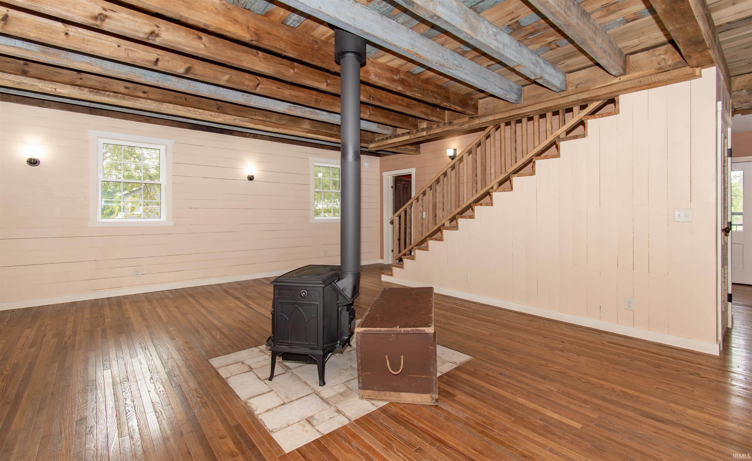 Open living area featuring a cast iron wood-burning stove on a stone hearth, exposed wood beam ceiling, hardwood flooring, and a natural wood staircase