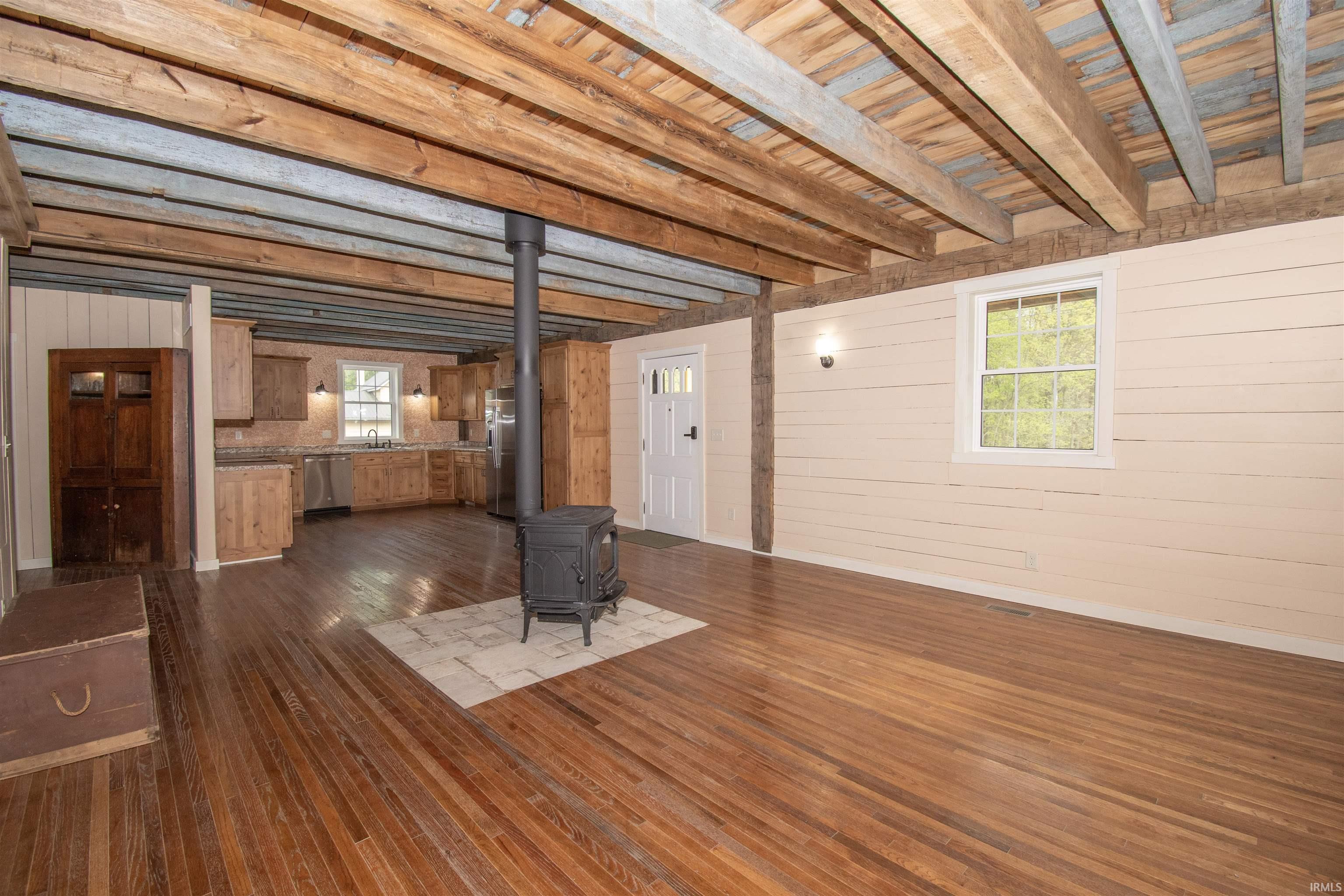 Open-concept living space featuring wood-finish flooring, exposed wood beam ceiling, and a central wood-burning stove on a tiled hearth