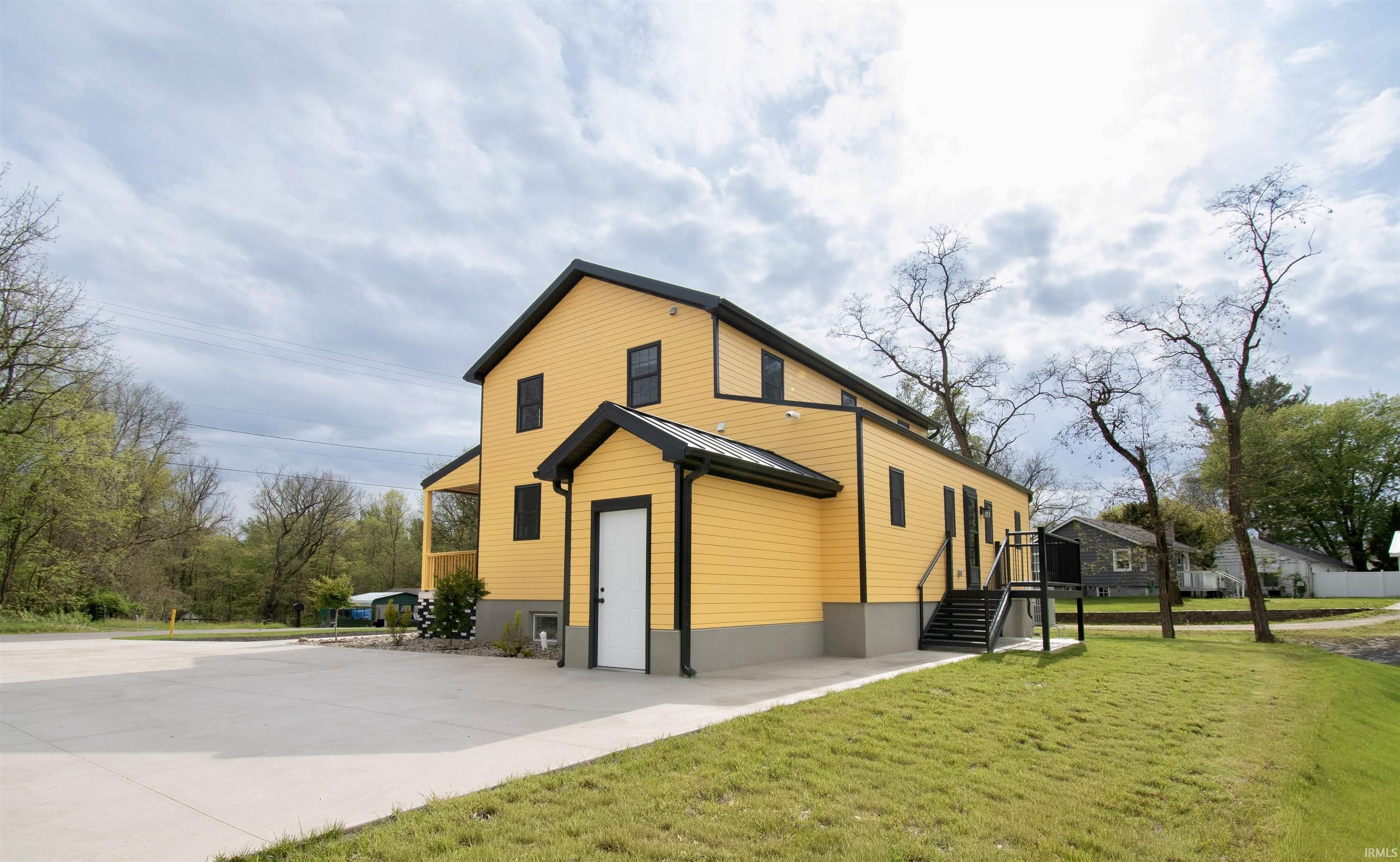 Two-story structure featuring yellow siding and black trim, a large paved parking area, a covered entrance, a separate side entrance with exterior stairs, and a sloped grassy area