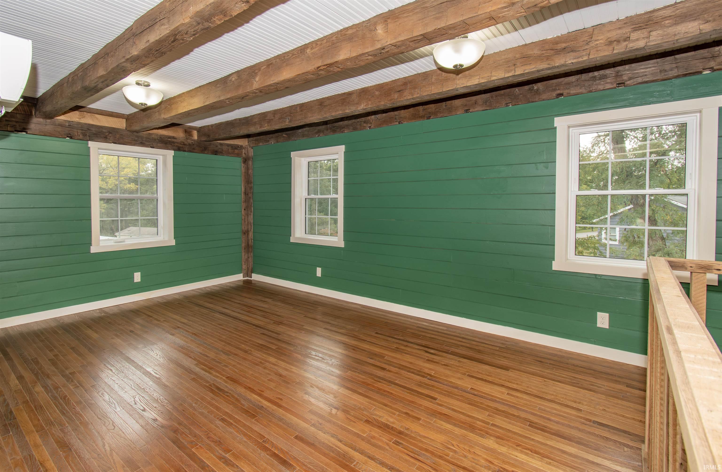 Room featuring wood-finish flooring, vibrant green horizontal plank walls, three double-hung windows with white trim, exposed ceiling beams, and a corrugated metal ceiling