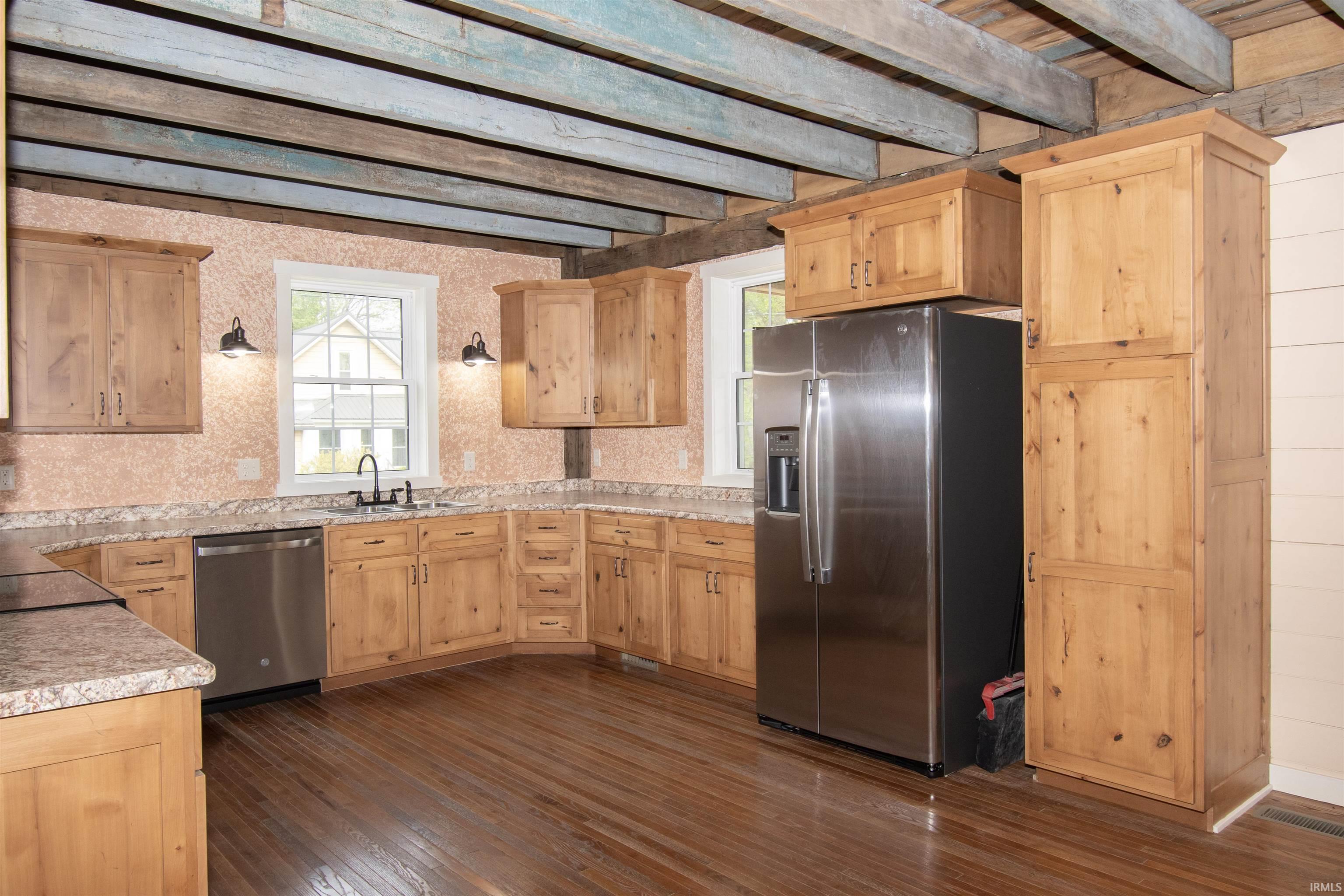 Kitchen featuring exposed ceiling beams, wood-finish flooring, light wood cabinetry, stone-look countertops, and stainless steel appliances