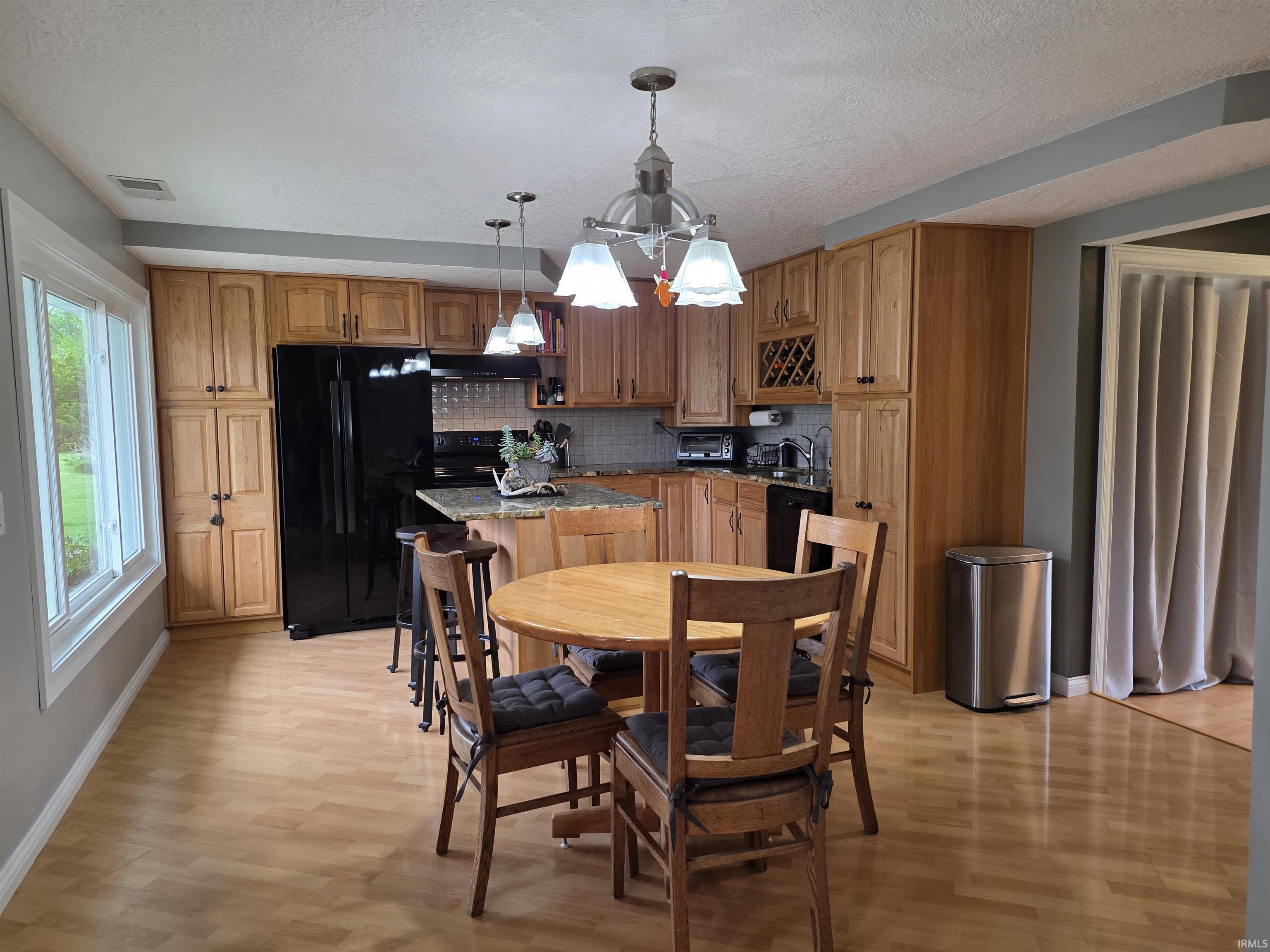 Spacious eat-in kitchen featuring wood-finish flooring, a large window, extensive wood cabinetry, a center island with granite-style countertop, and black appliances