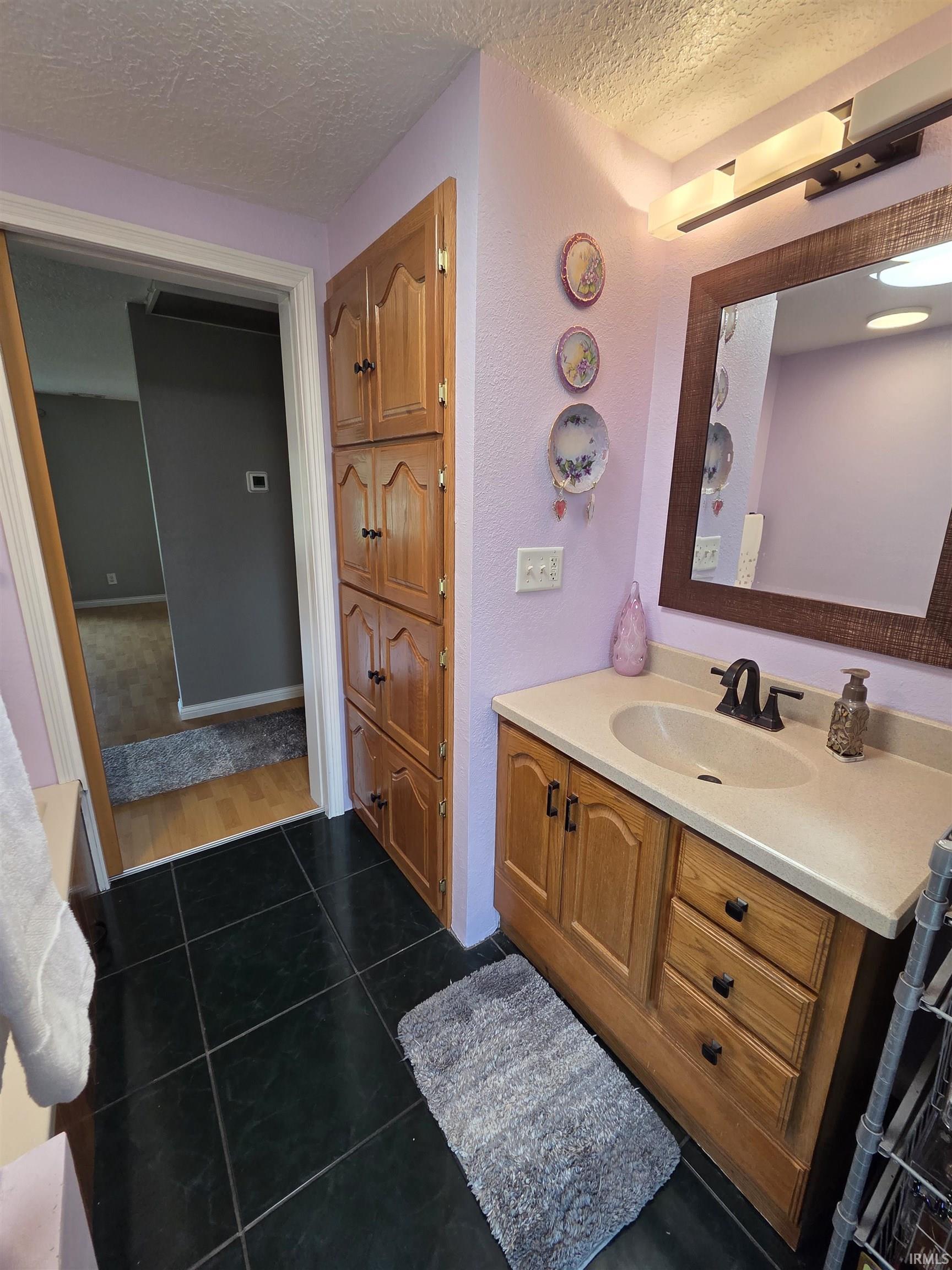 Bathroom featuring dark square floor tiles, a light wood-finish vanity with an integrated sink and light countertop, and a matching full-height storage cabinet