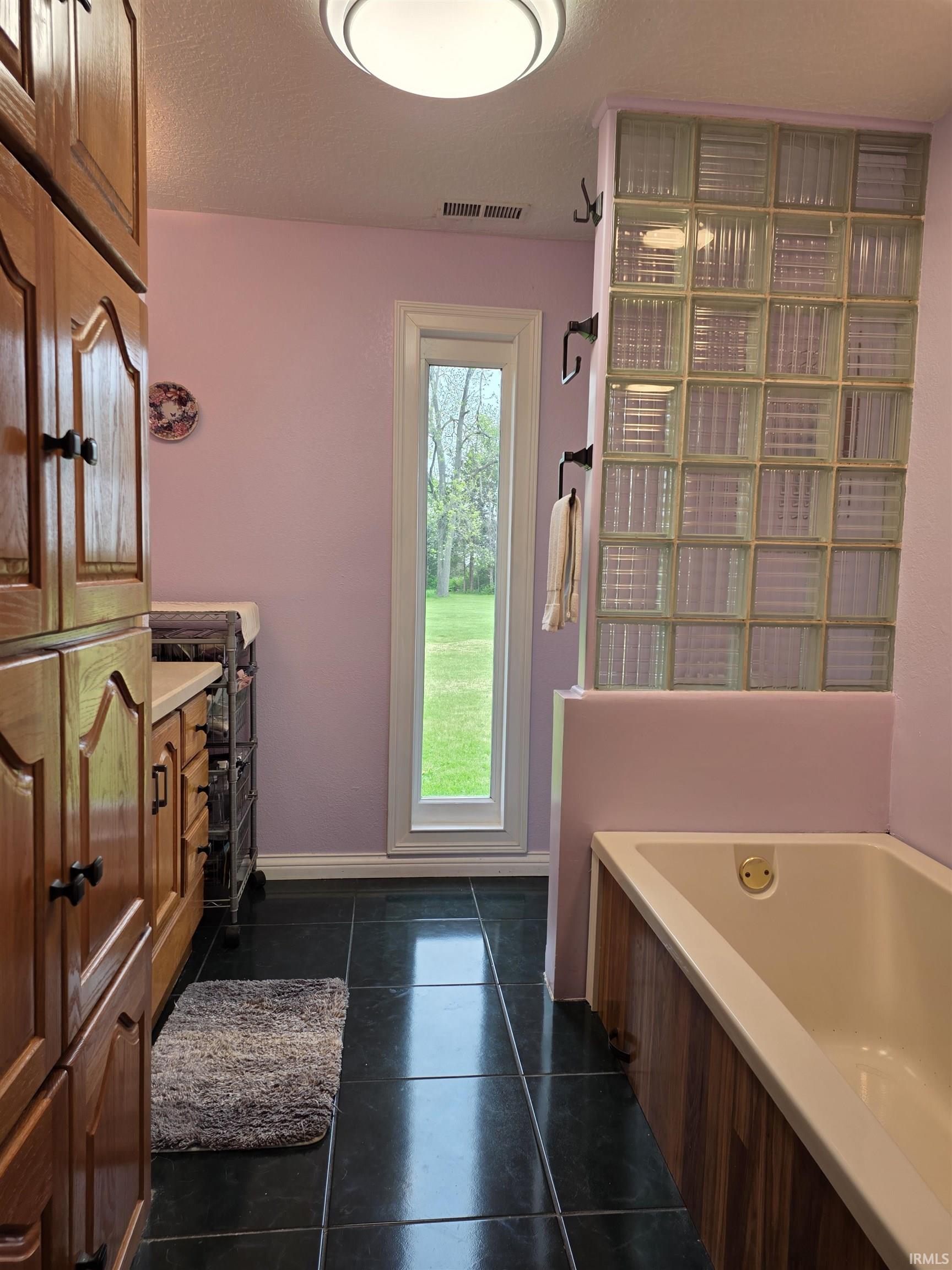 Bathroom featuring dark tile flooring, a built-in soaking tub with wood-finish paneling, and a glass block privacy wall