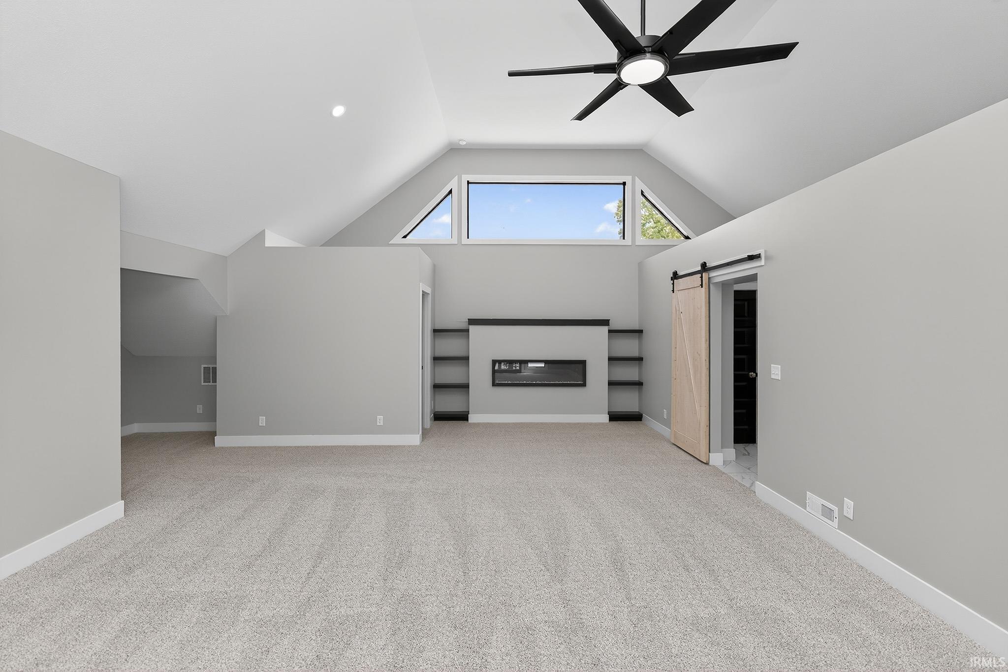 Spacious room featuring vaulted ceilings with a contemporary ceiling fan, multiple windows, plush carpet flooring, a built-in electric fireplace with shelving, and a barn door entry