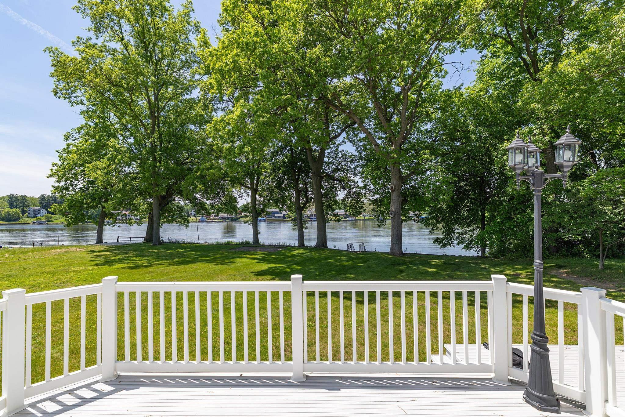 Deck with white railing and lamppost overlooking a grassy area and a body of water lined with mature trees