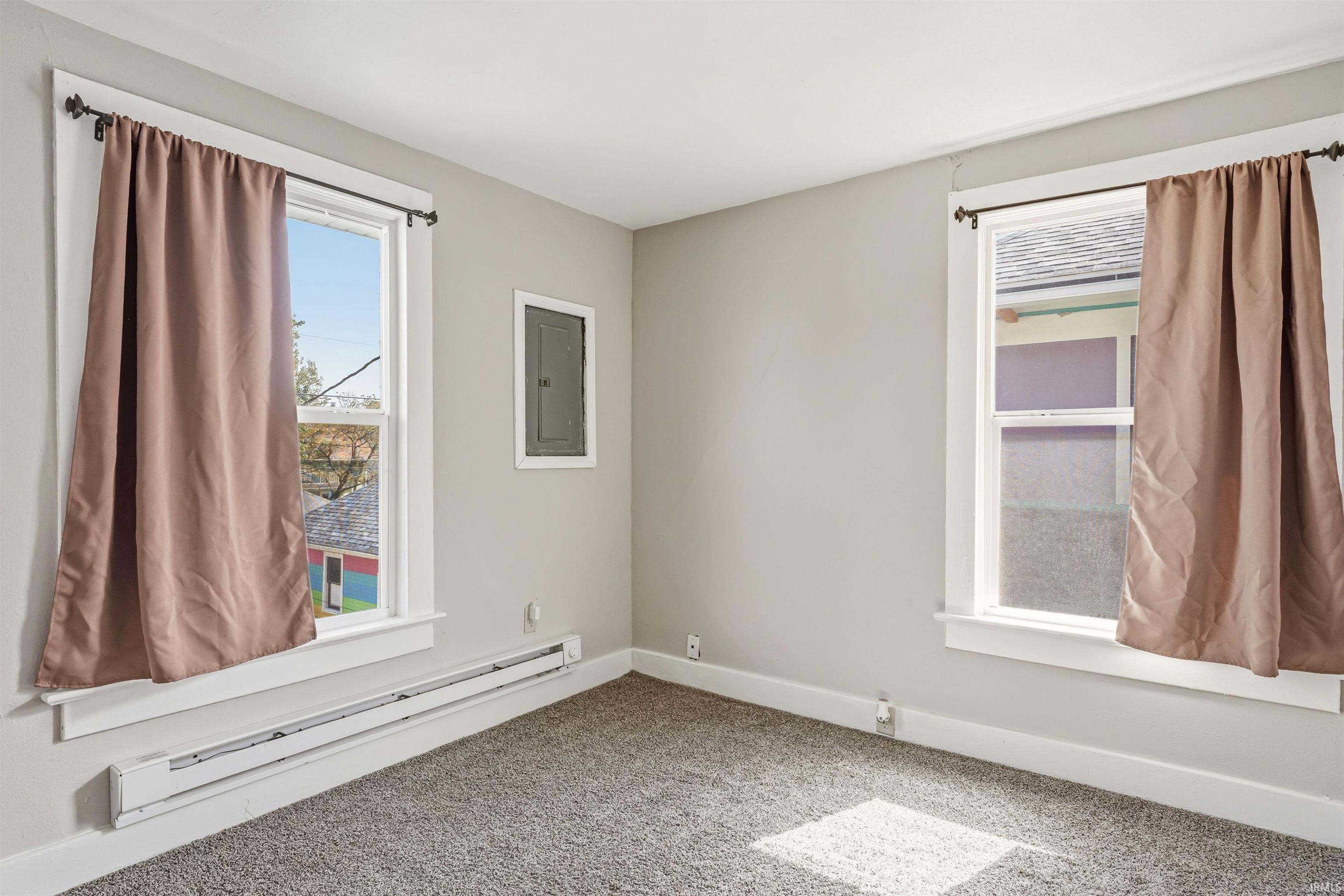 Carpeted interior space featuring two large windows with white trim, baseboard heating, and light gray painted walls