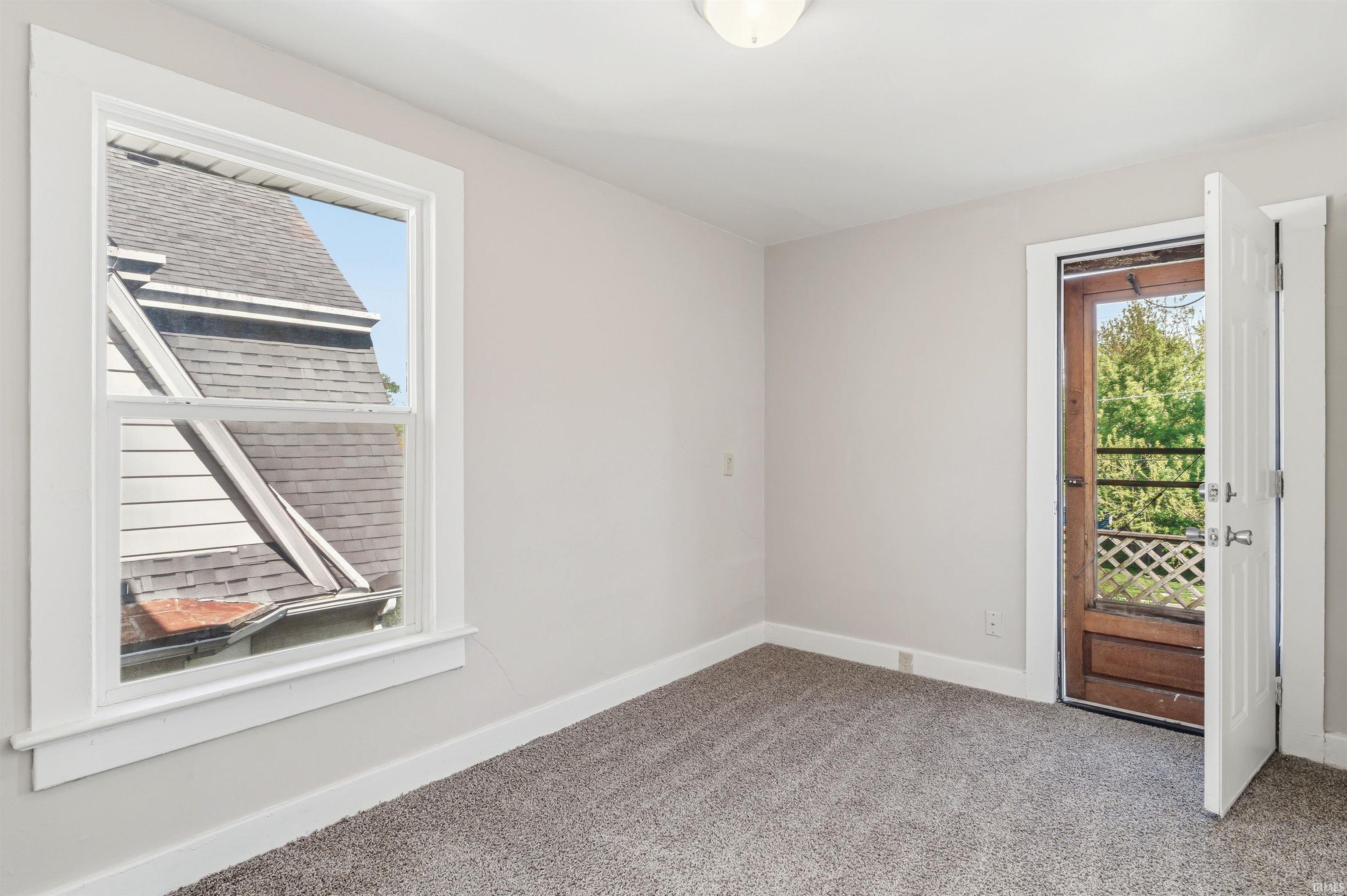 Bright room featuring light grey walls, grey carpet, a white framed window, and a white interior door opening to a dark wood exterior door with a lattice screen