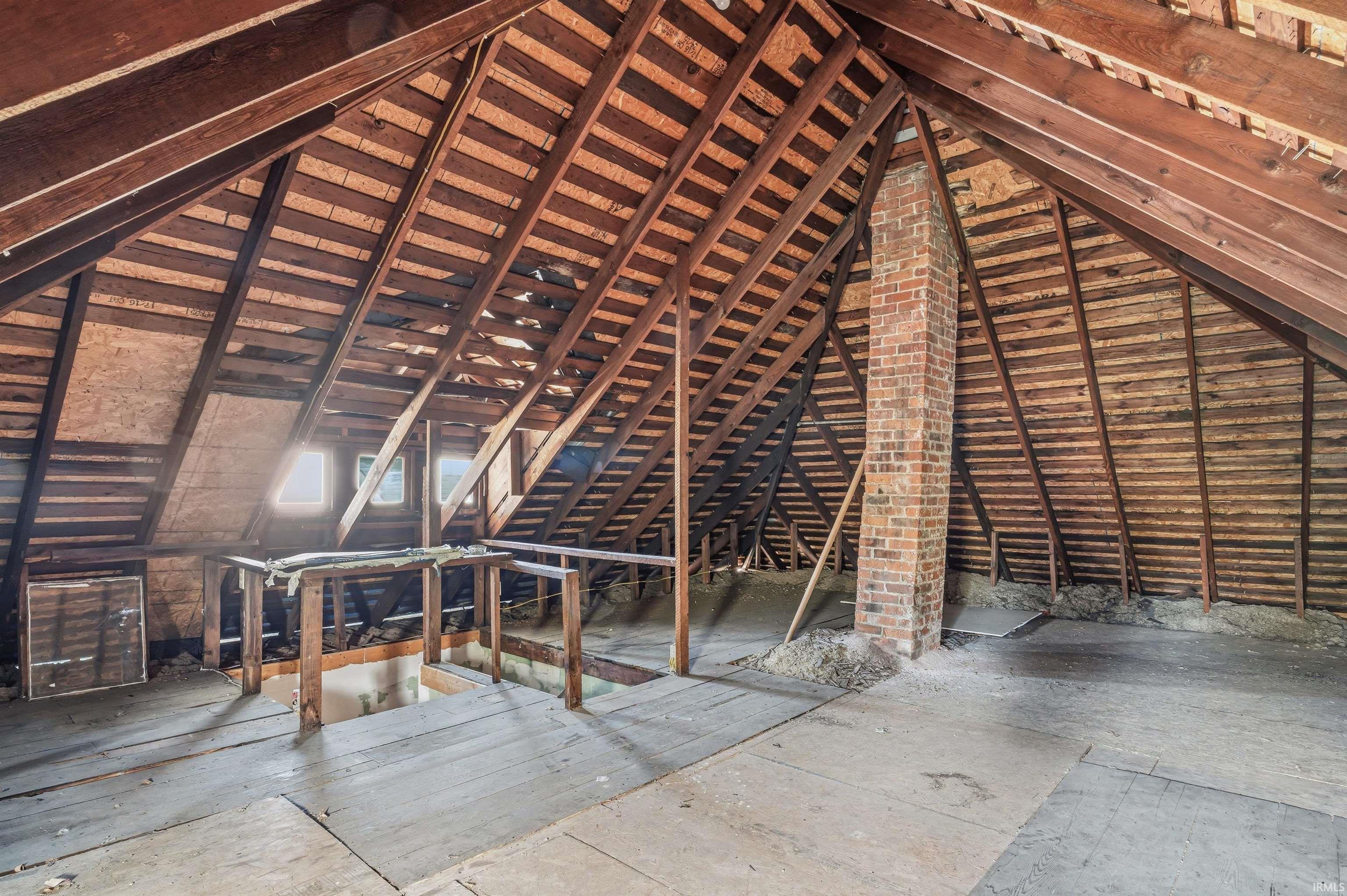 Spacious attic area featuring exposed wood beams and joists, a prominent brick chimney, two visible windows providing natural light, and ample floor space