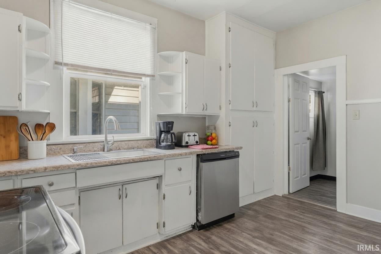 Kitchen featuring extensive white cabinetry, a stainless steel dishwasher, a single basin sink with a gooseneck faucet, light-toned countertops, and wood-finish flooring