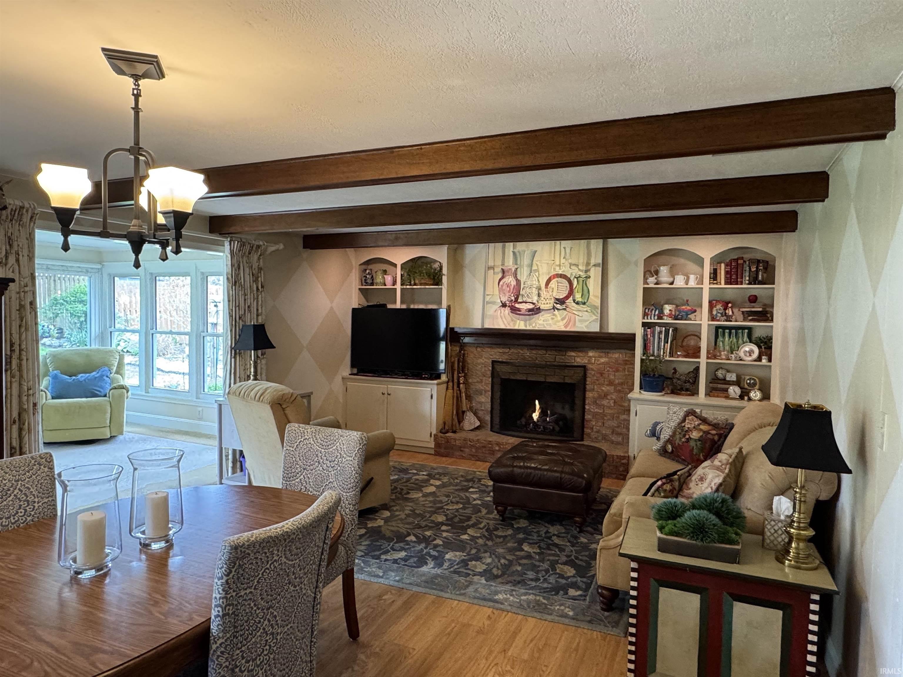 Spacious living area featuring exposed ceiling beams, a brick-surround fireplace, custom built-in shelving, and wood-finish flooring