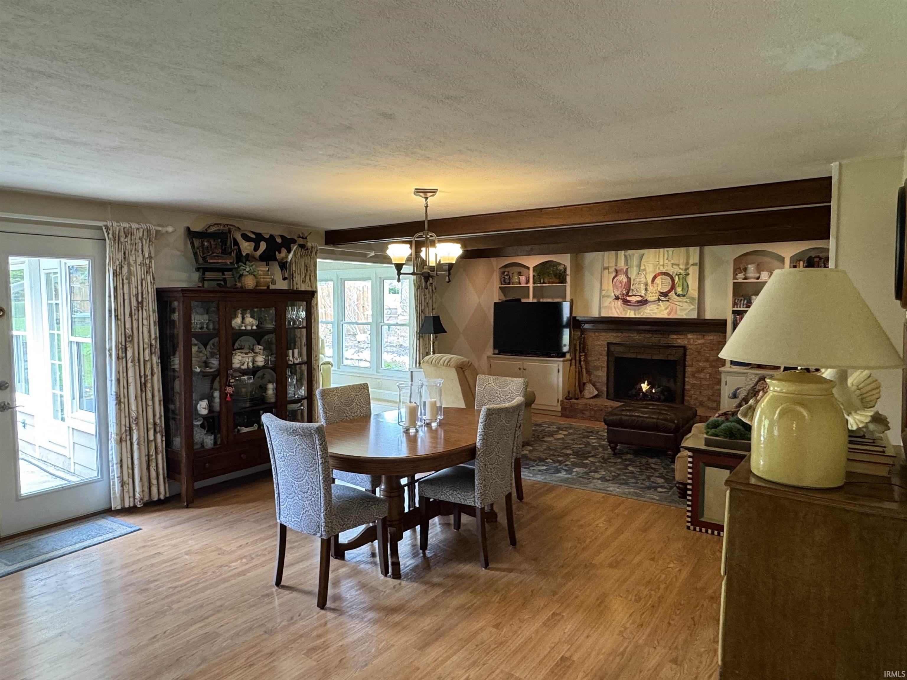 Wood-finish flooring throughout, featuring a brick-surround fireplace with built-in shelving, exposed ceiling beams, and a ceiling-mounted light fixture