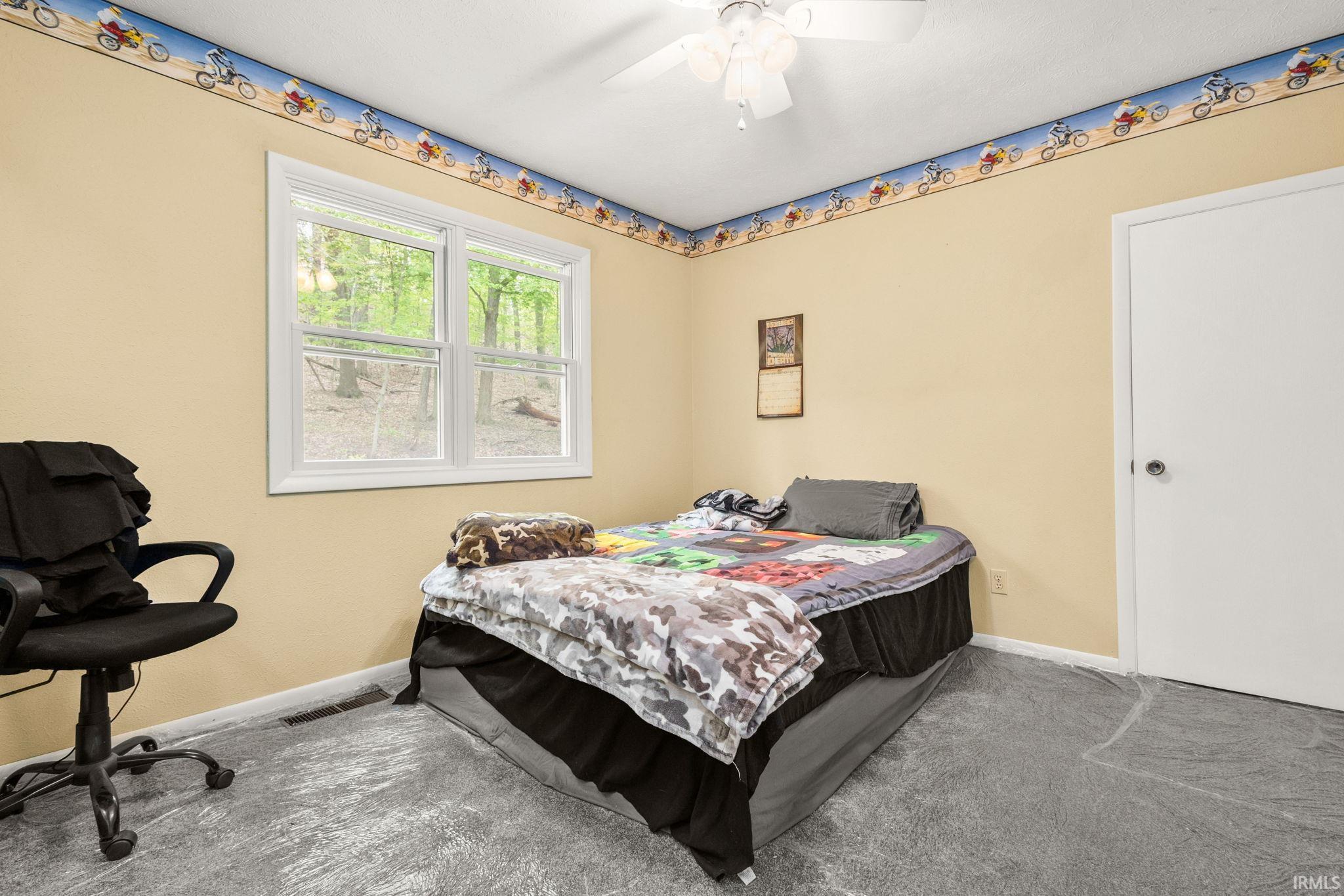 Room featuring light-toned walls, grey patterned carpeting, a white ceiling fan, and a double window with a wooded backdrop