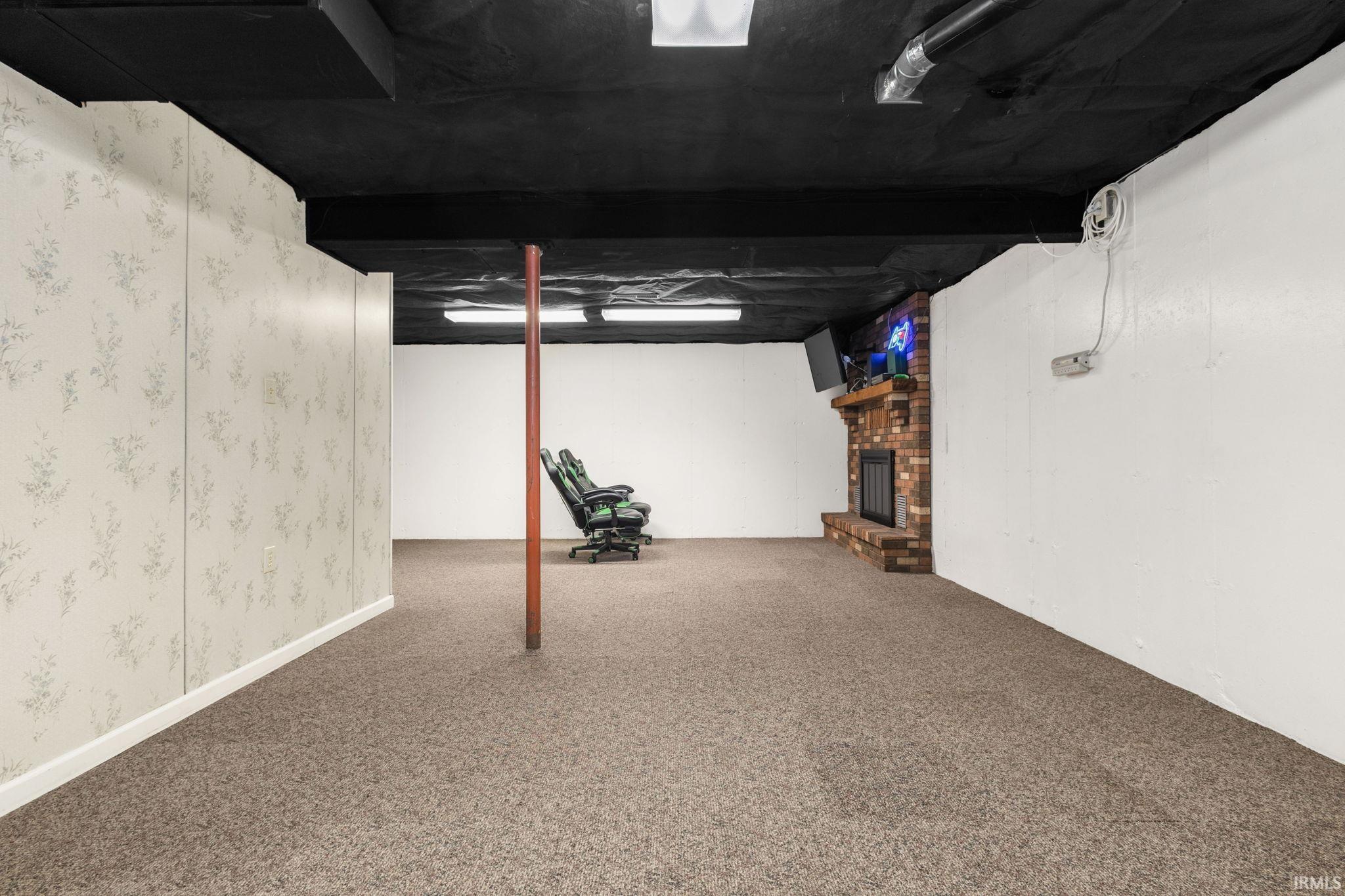 Spacious lower-level area with carpet flooring, a brick fireplace with a wood mantle, and exposed black ceiling joists