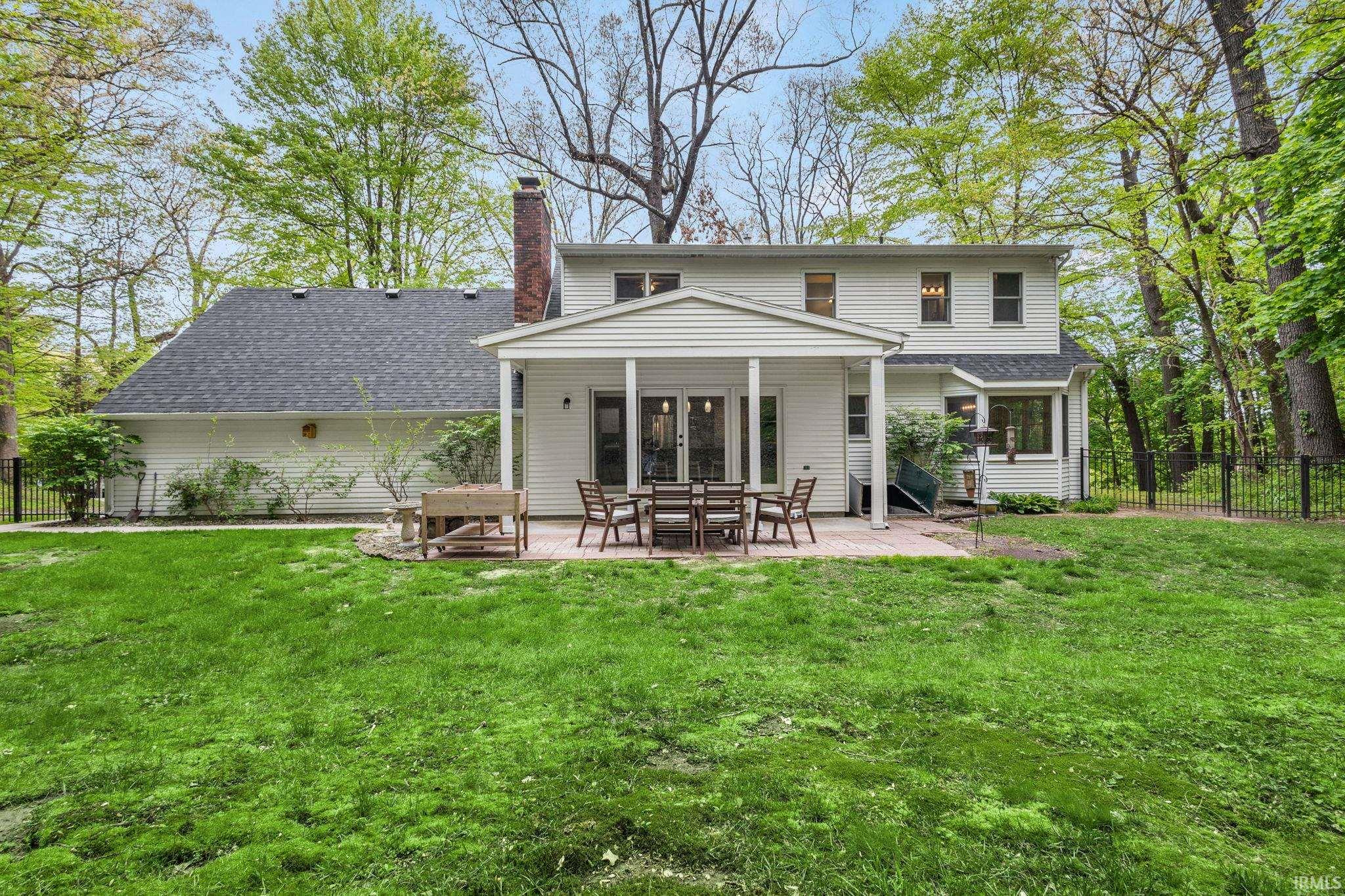 Expansive rear yard featuring a covered patio with a paved surface, mature trees, and a black metal fence