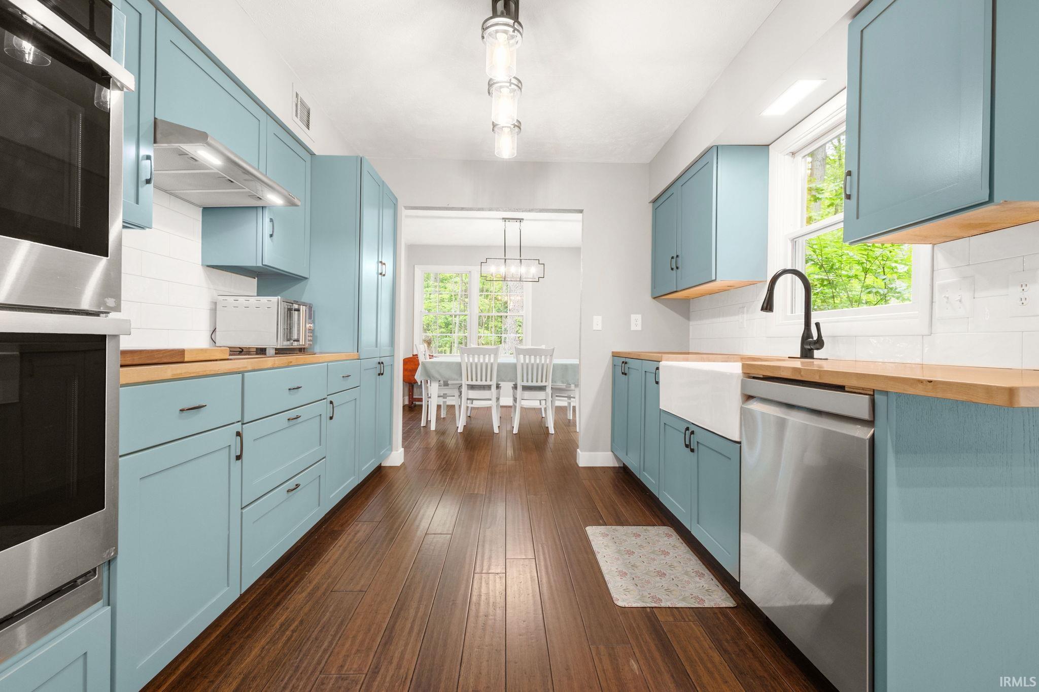 Galley kitchen featuring wood-finish flooring, light blue cabinetry with dark hardware, butcher block countertops, a white farmhouse sink, and stainless steel appliances