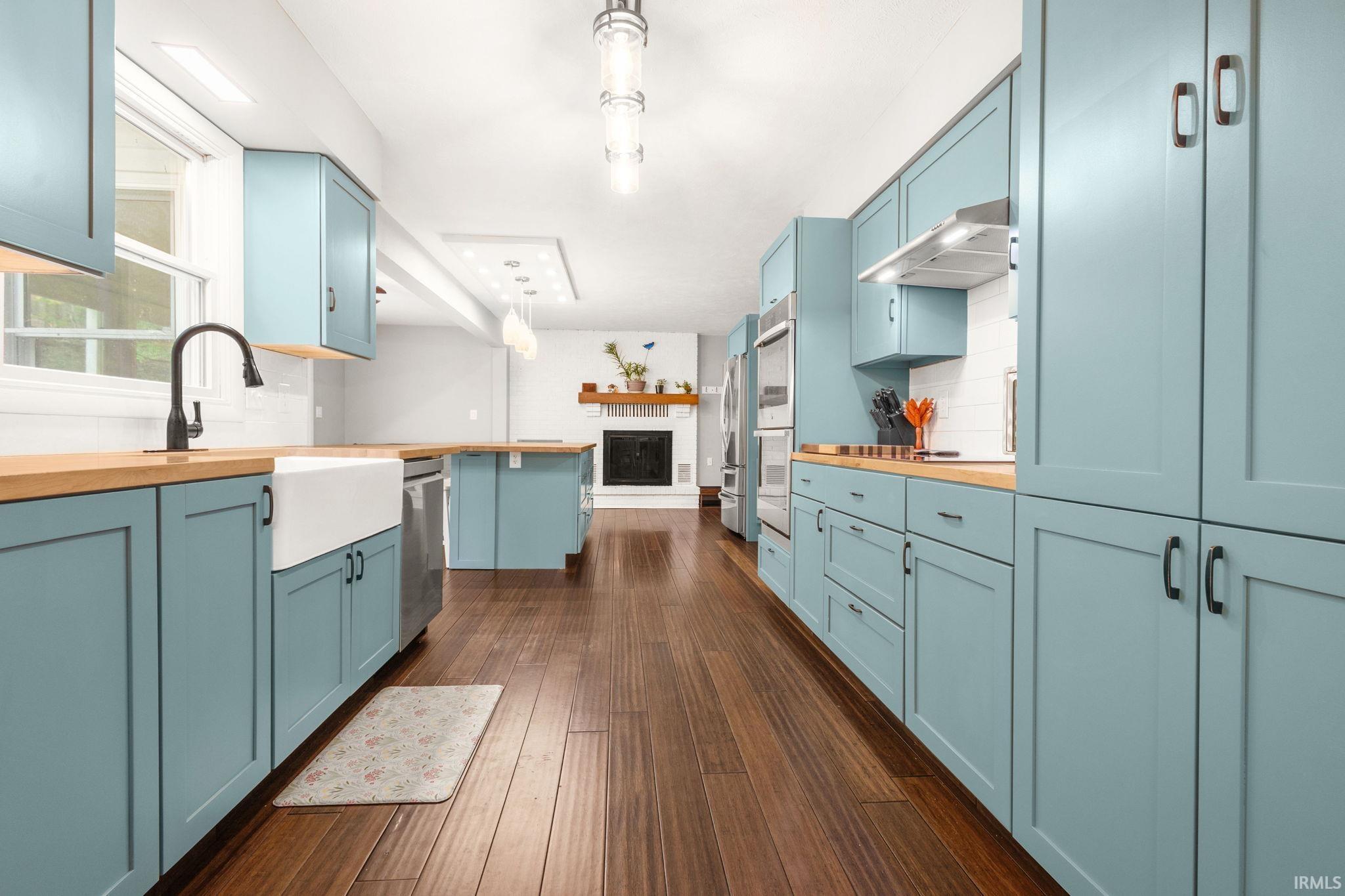Spacious kitchen featuring light blue cabinetry, butcher block countertops, a farm sink with a matte black faucet, stainless steel appliances, and wood-finish flooring