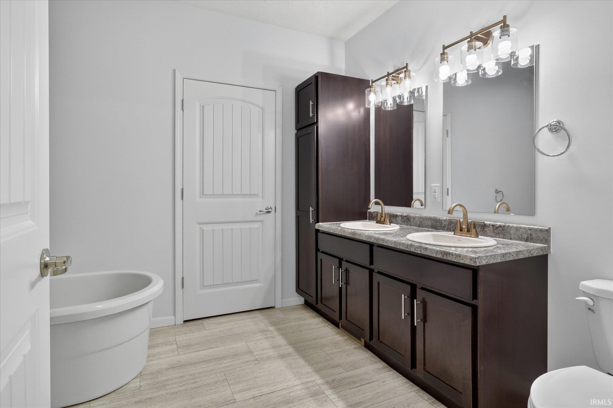 Bathroom featuring a dual vanity with dark wood cabinetry, a tall built-in storage cabinet, brushed brass fixtures, and a white soaking tub