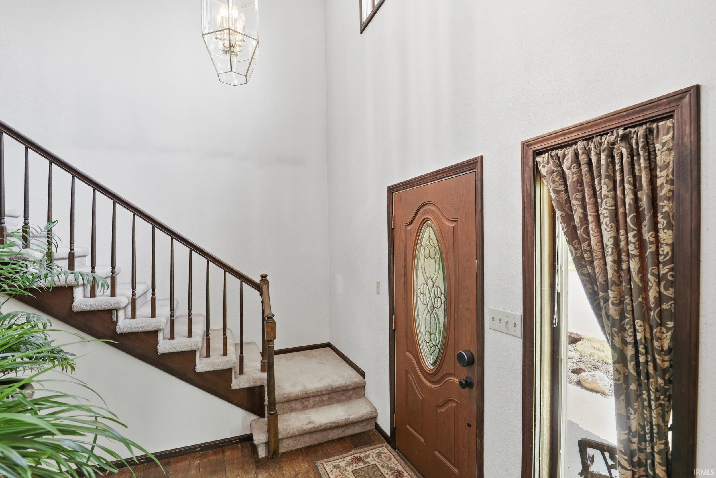 Double-height foyer featuring wood-finish flooring, a carpeted staircase with wood banister, an ornate glass-insert entry door, and a geometric pendant light fixture