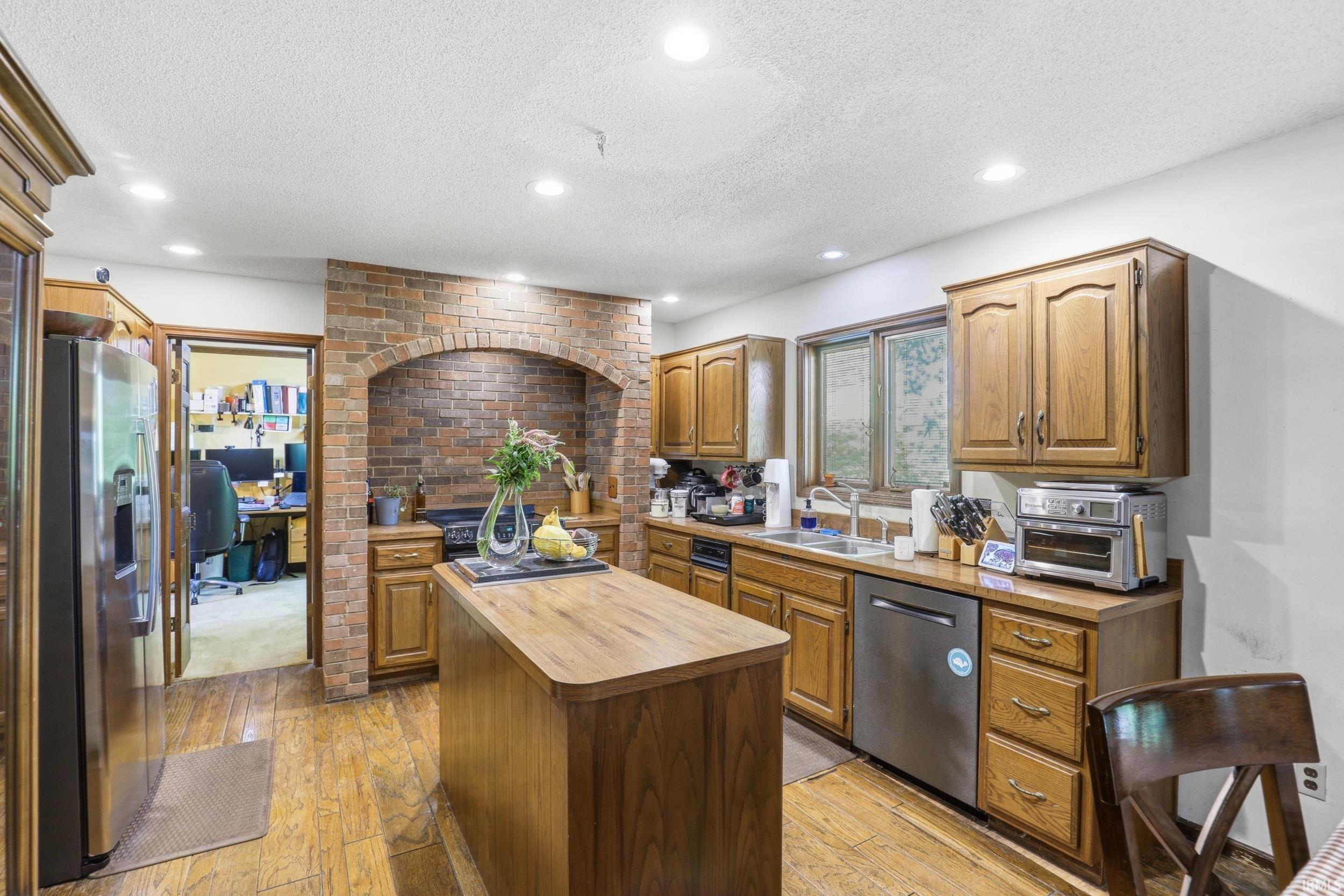 Kitchen featuring a brick accent wall with an arched alcove, wood-finish flooring, recessed lighting, wood cabinetry, and a stainless steel refrigerator
