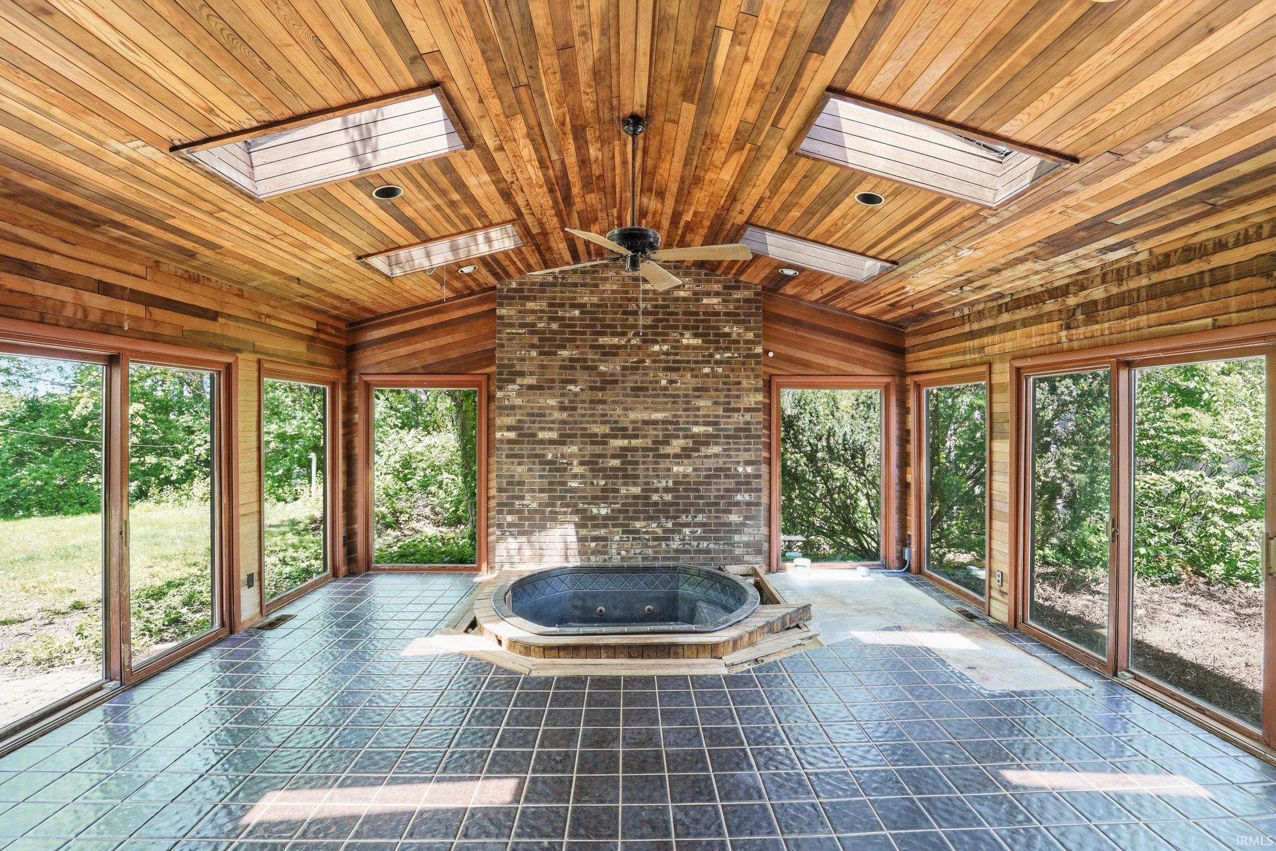 Sunroom featuring a built-in hot tub, dark tiled flooring, a brick accent wall, and a wood-paneled ceiling with skylights and a ceiling fan