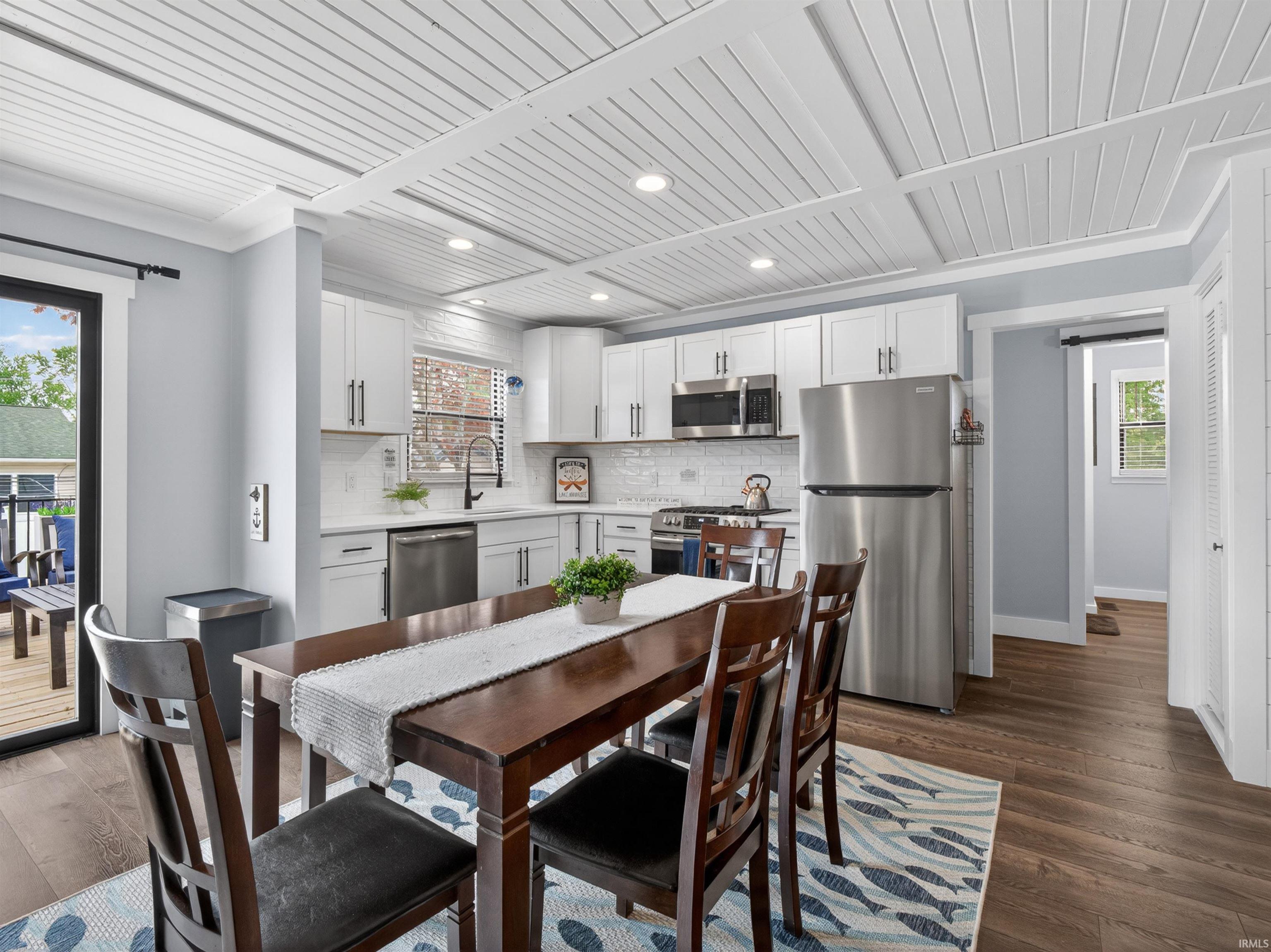 Open-concept kitchen and dining area featuring white shaker cabinetry, stainless steel appliances, wood-finish flooring, a white subway tile backsplash, and a coffered plank ceiling