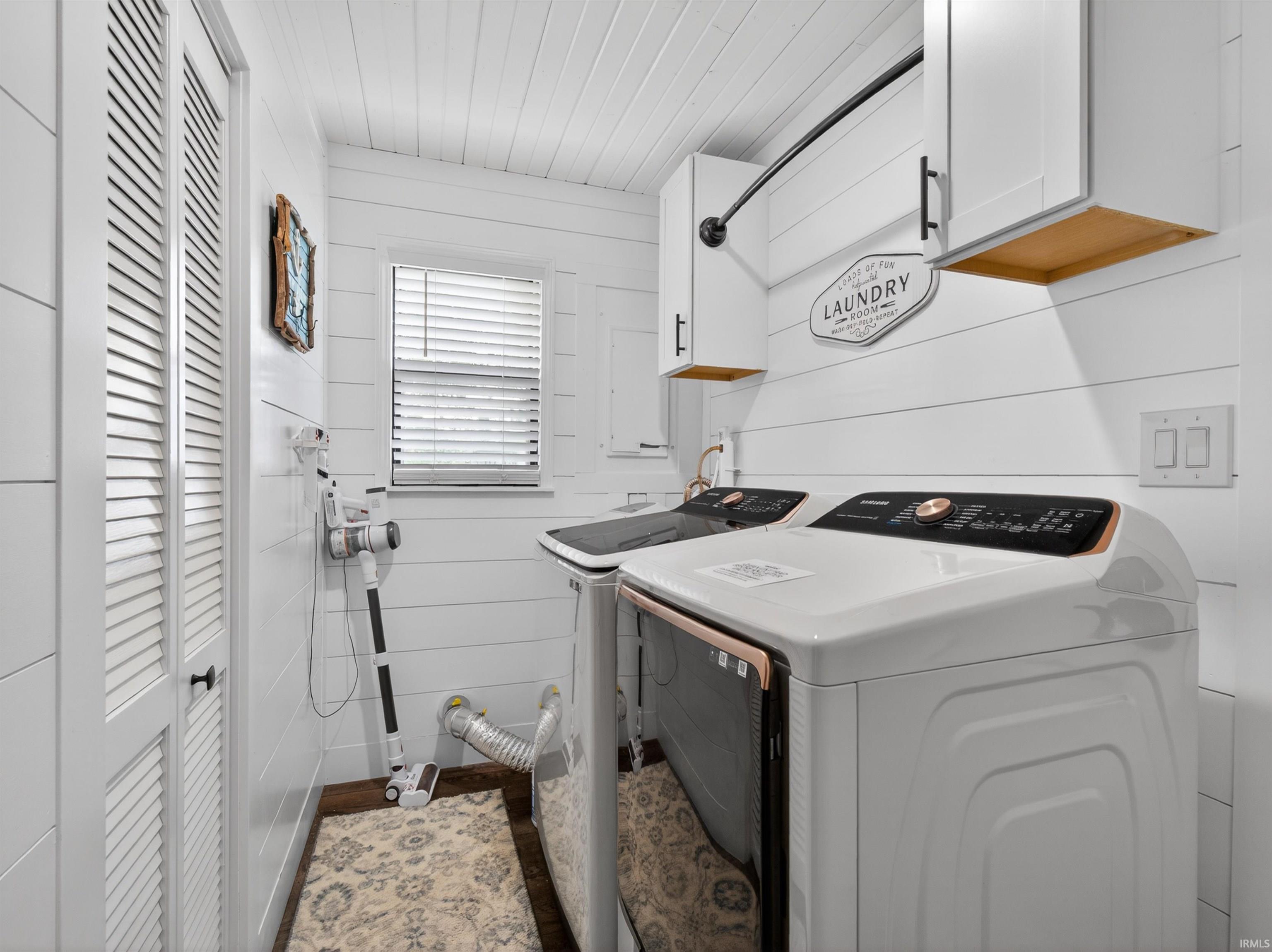 Laundry area with shiplap walls, wood-finish flooring, louvered bi-fold doors, and upper cabinetry with black hardware