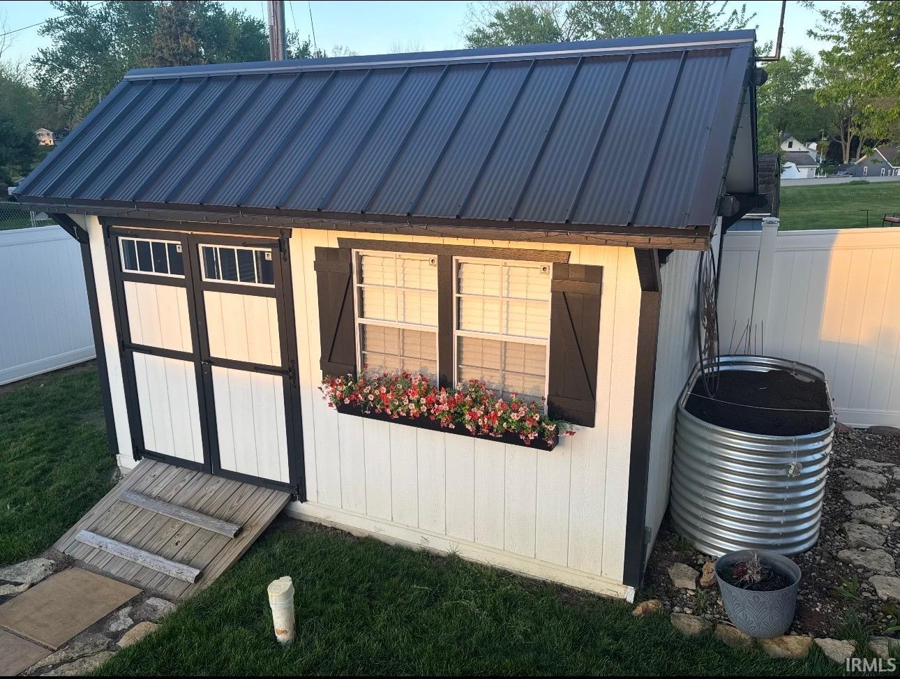White storage shed featuring a black metal roof, double entry doors with transom windows, and a window box with plantings
