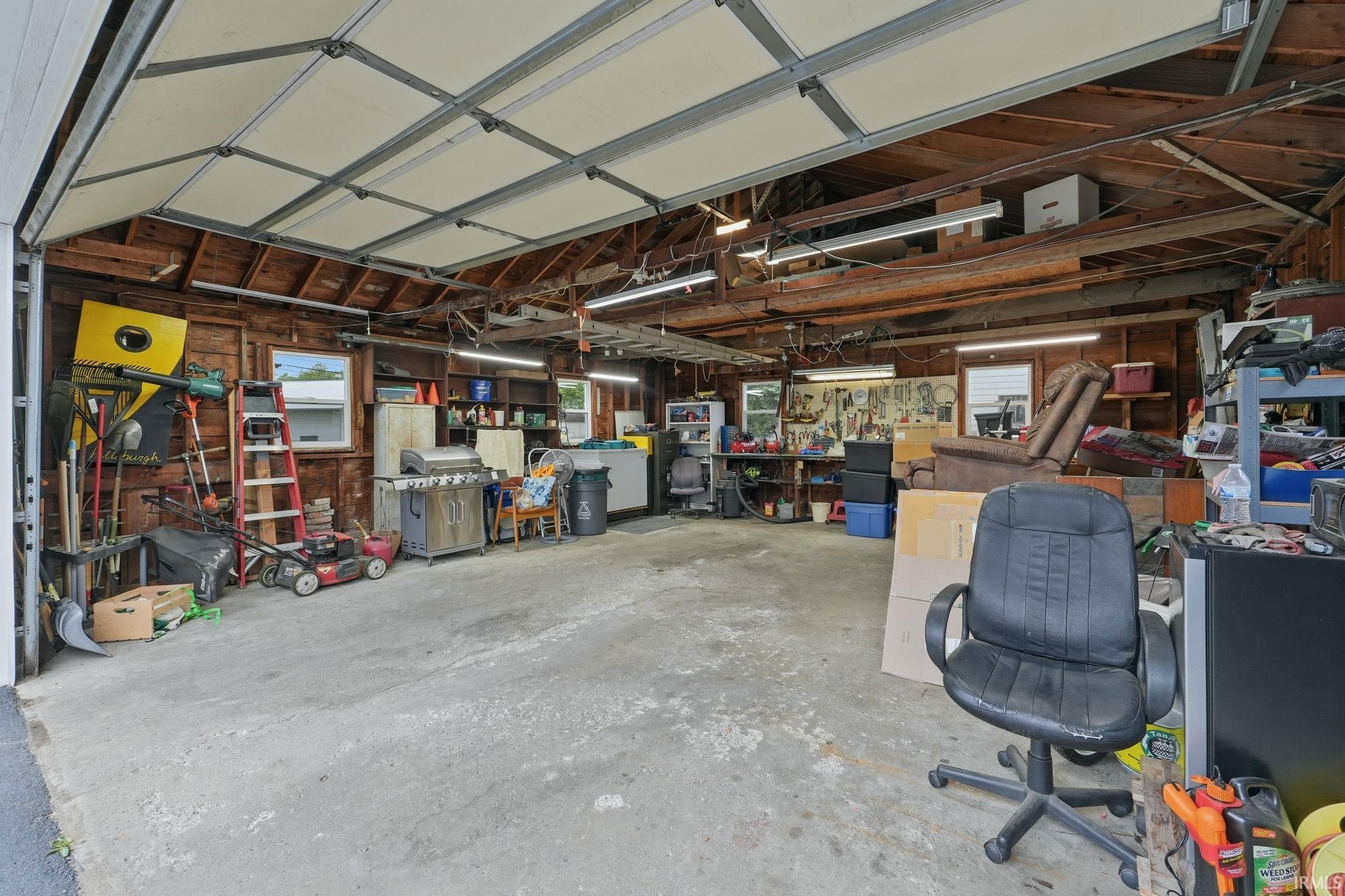 Spacious garage featuring an overhead door, concrete flooring, exposed wood beam ceiling, multiple windows, and extensive built-in shelving