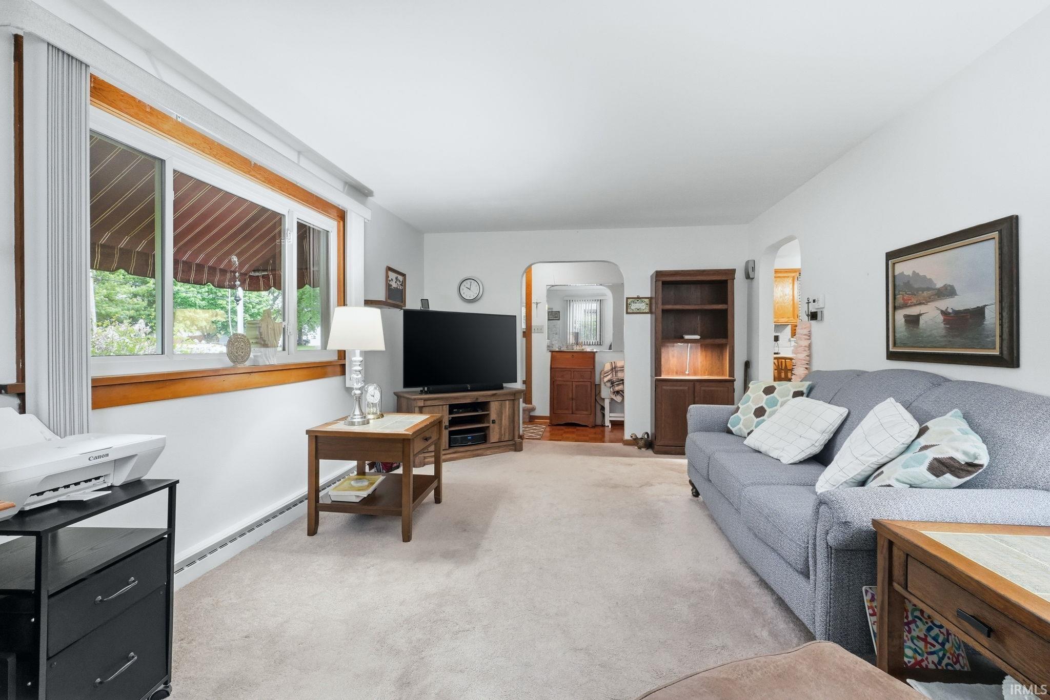 Spacious living area featuring light-colored carpeting, a large picture window with wood trim, and an arched doorway leading to an additional area