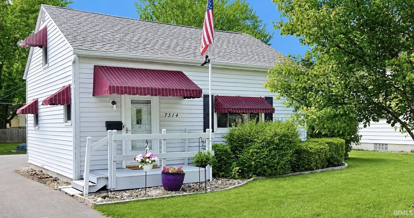 White siding exterior with a shingled roof, featuring burgundy striped window awnings, a front porch with white railing, and established perimeter landscaping