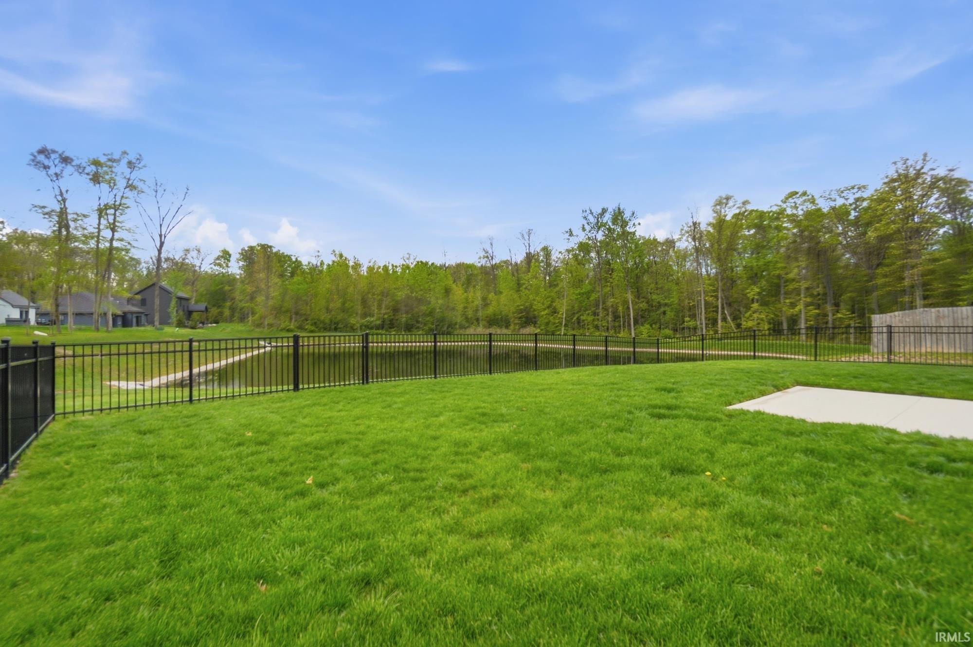 Expansive green lawn featuring a black wrought iron fence bordering a pond with a wooded perimeter