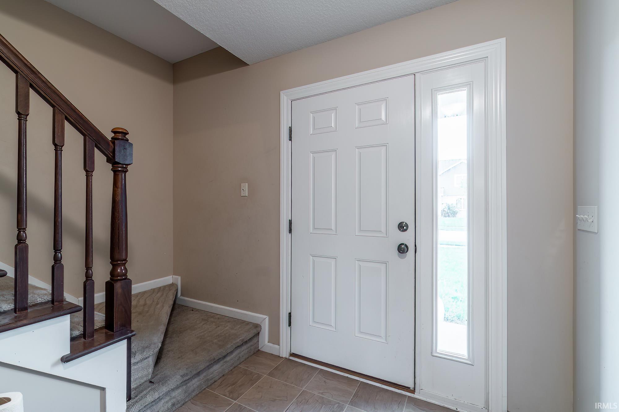 Entryway featuring a paneled white door with a clear glass sidelight, tile flooring, and a carpeted staircase with wood banister