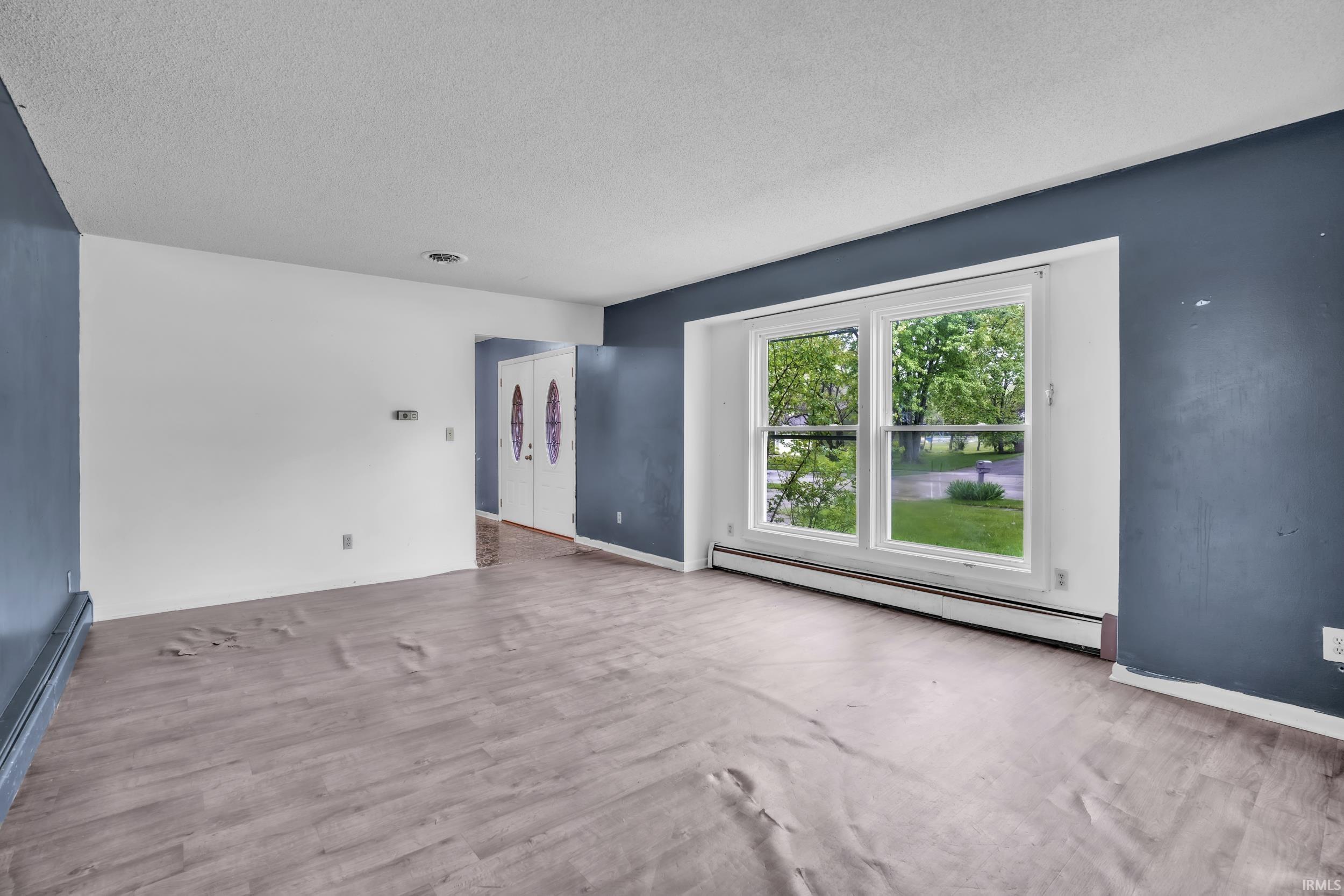 Spacious room with light wood-finish flooring, a large window providing ample natural light, and a double entry door with decorative glass inserts