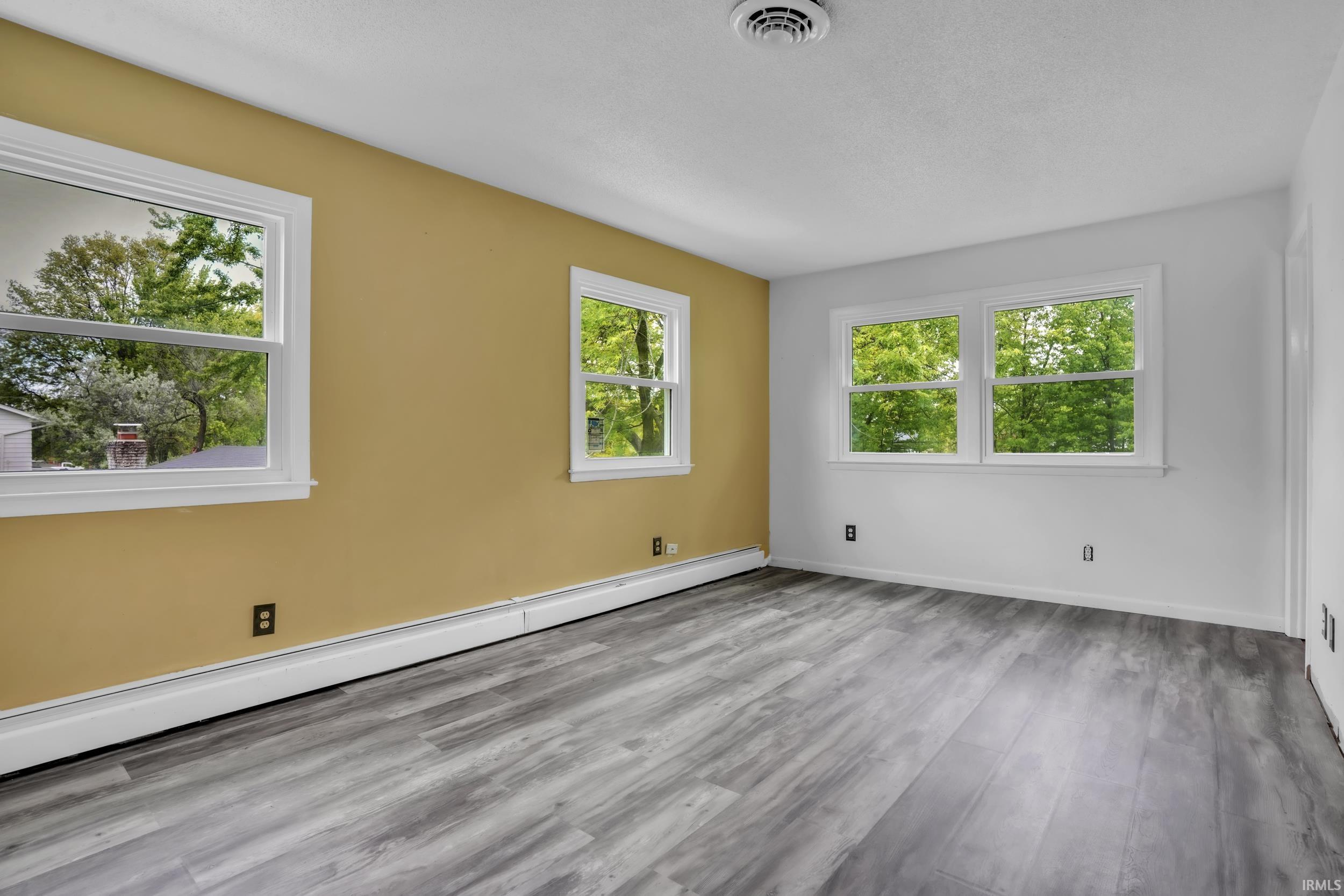 Spacious room featuring light-toned wood-finish flooring, multiple white-framed windows, and baseboard heating