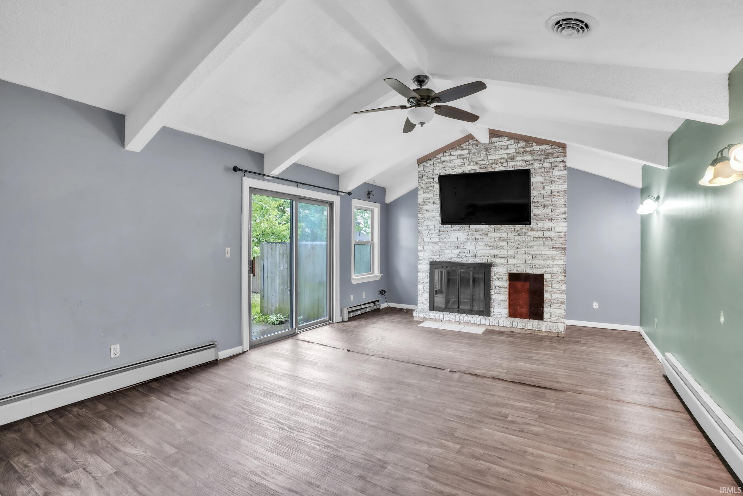 Living area featuring wood-finish flooring, exposed beam vaulted ceilings, a stone-clad fireplace, ceiling fan, and sliding glass door access to the exterior